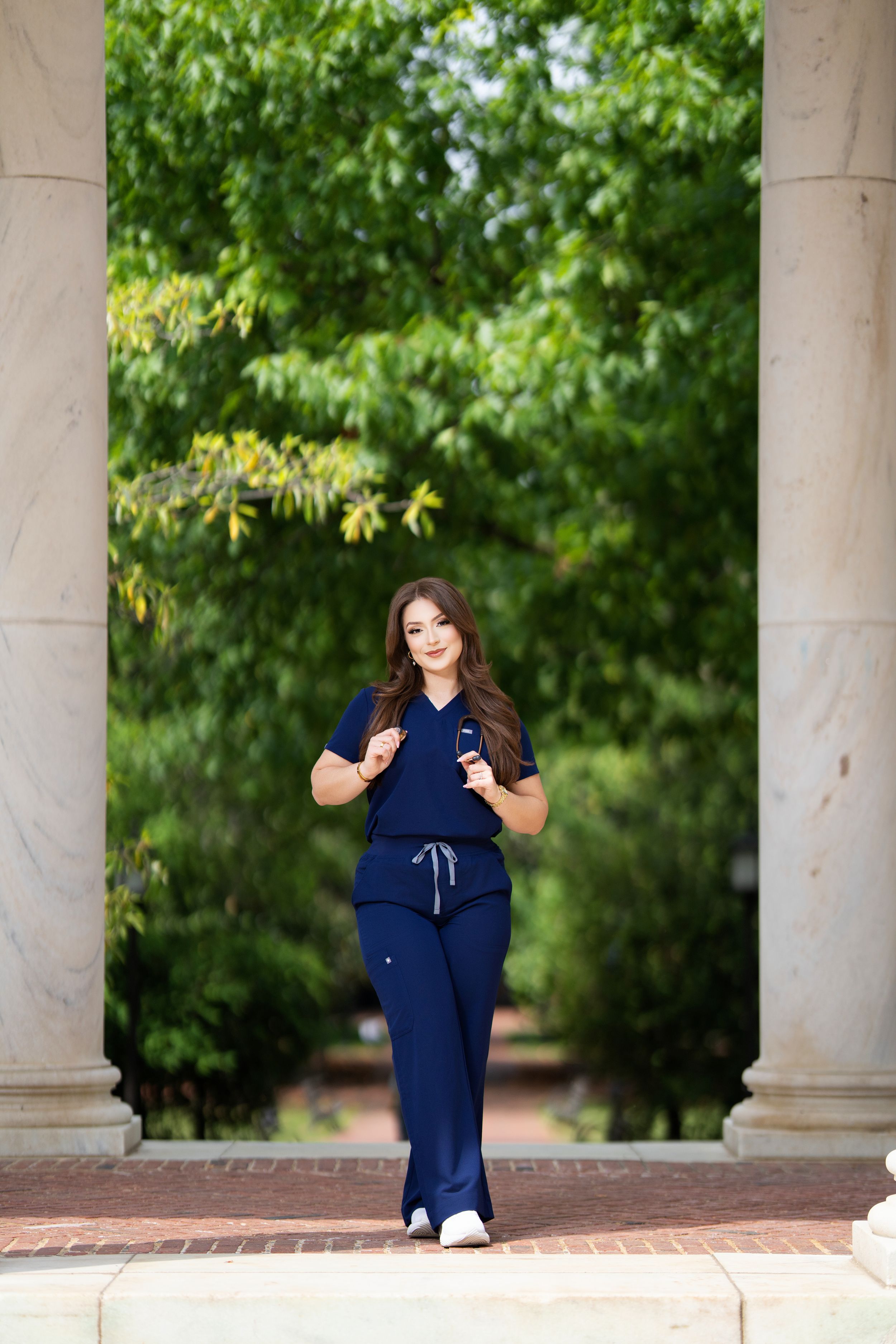 Young woman in navy scrubs walks between marble columns in green park.