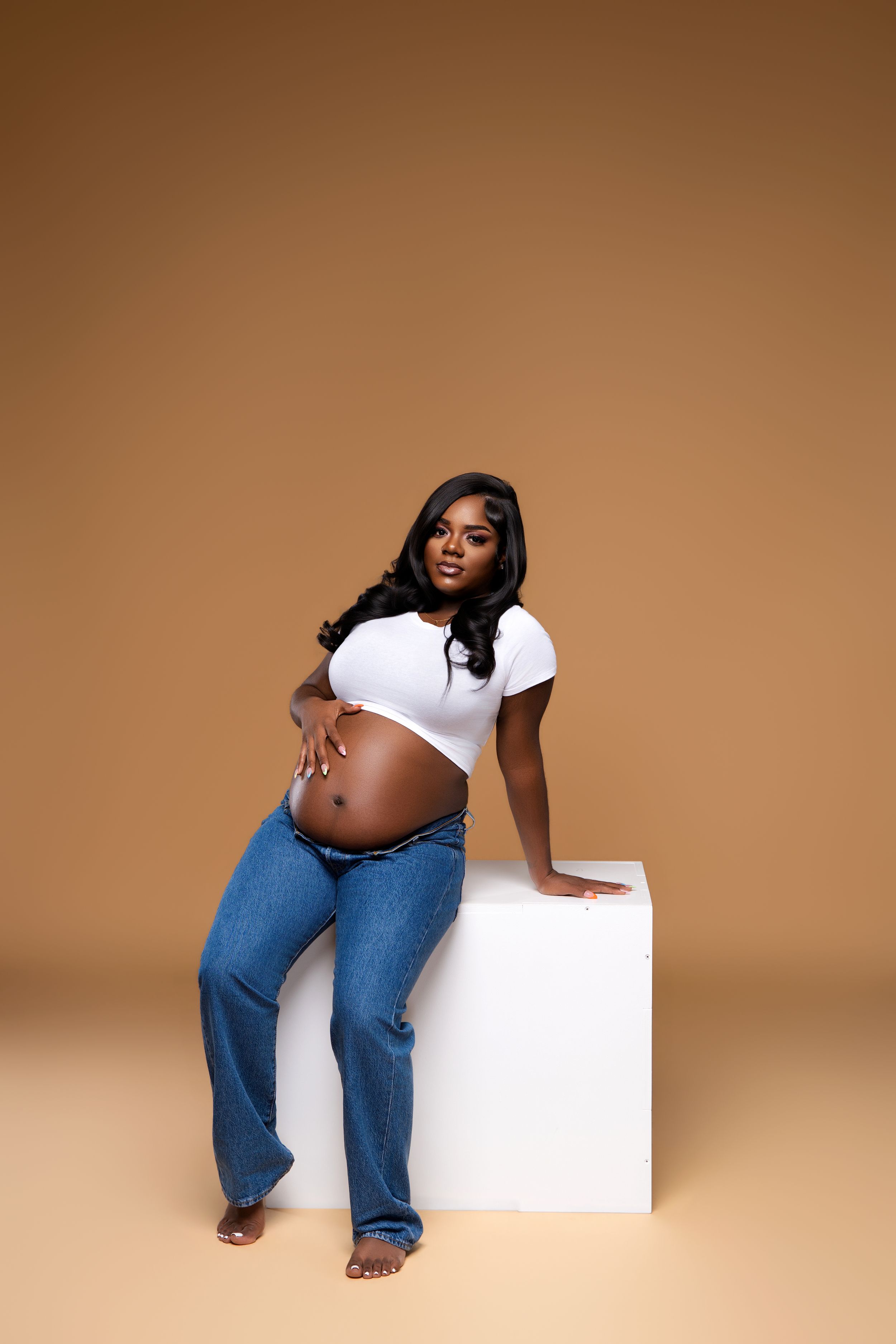 Pregnant woman in white crop top and jeans leans on white cube against warm beige background.