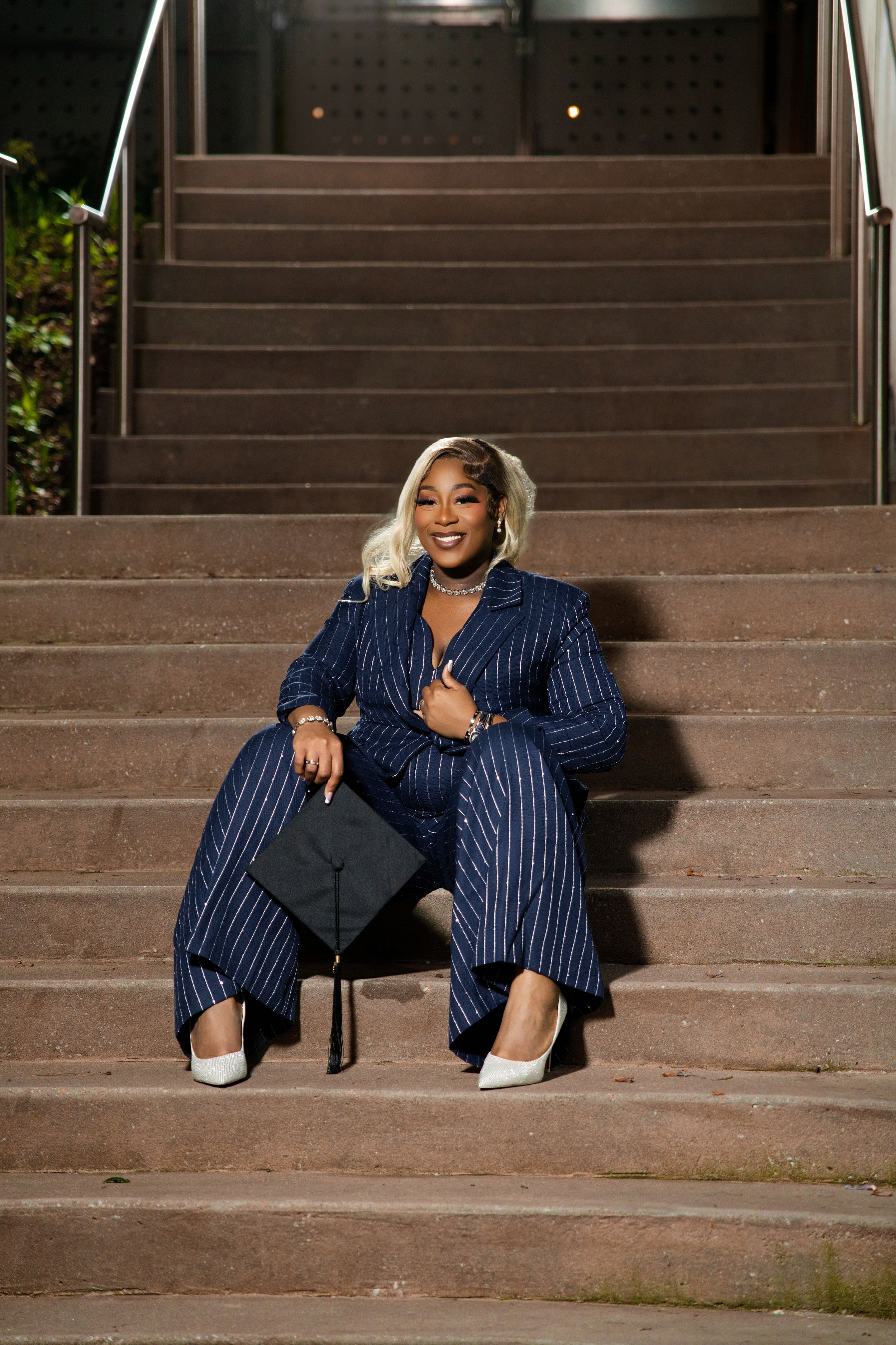 Black woman in navy pinstripe suit holds graduation cap while sitting on outdoor steps at night.