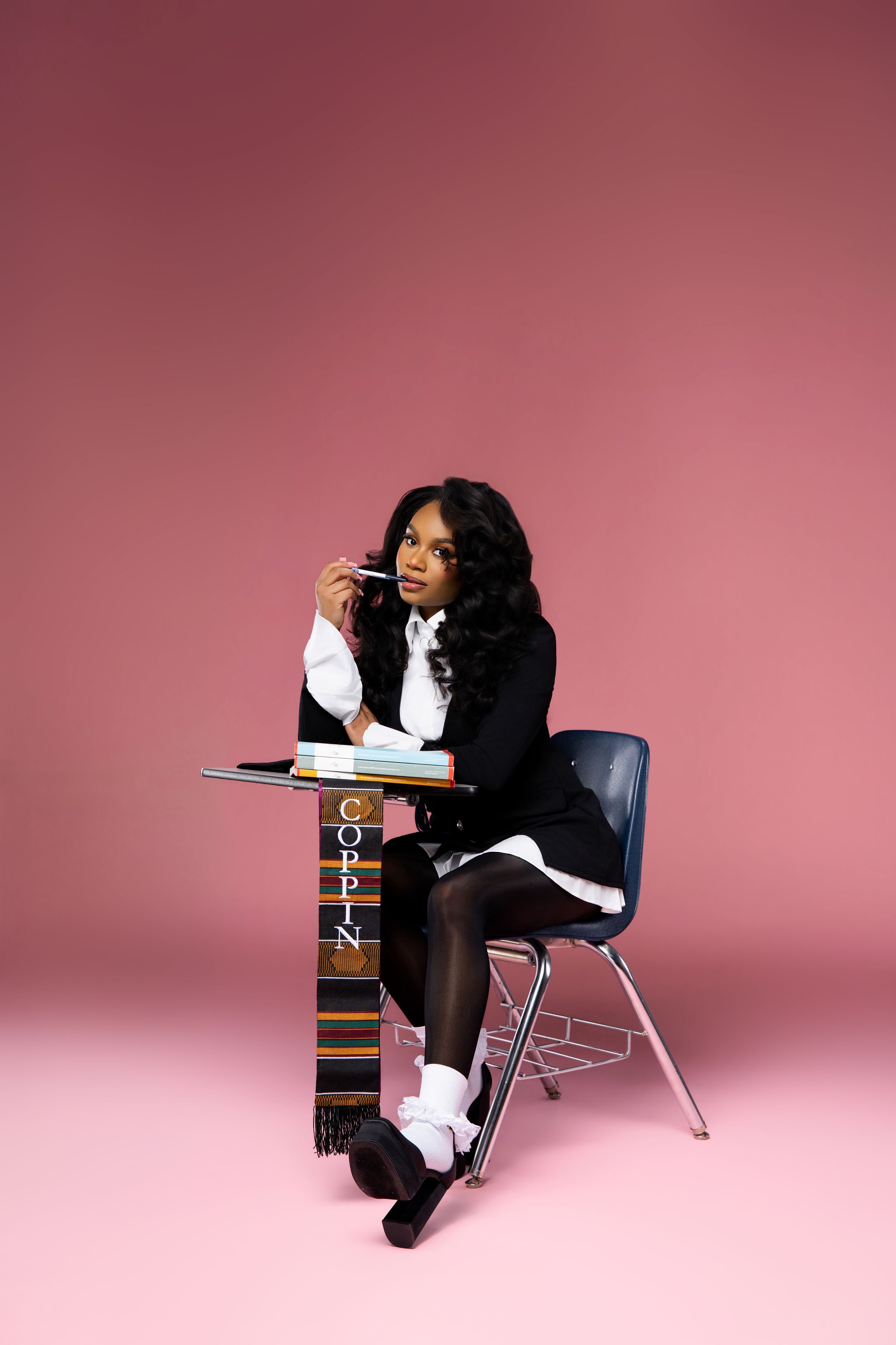 Black woman in school uniform sits at desk with Coppin stole and pencil on pink background.