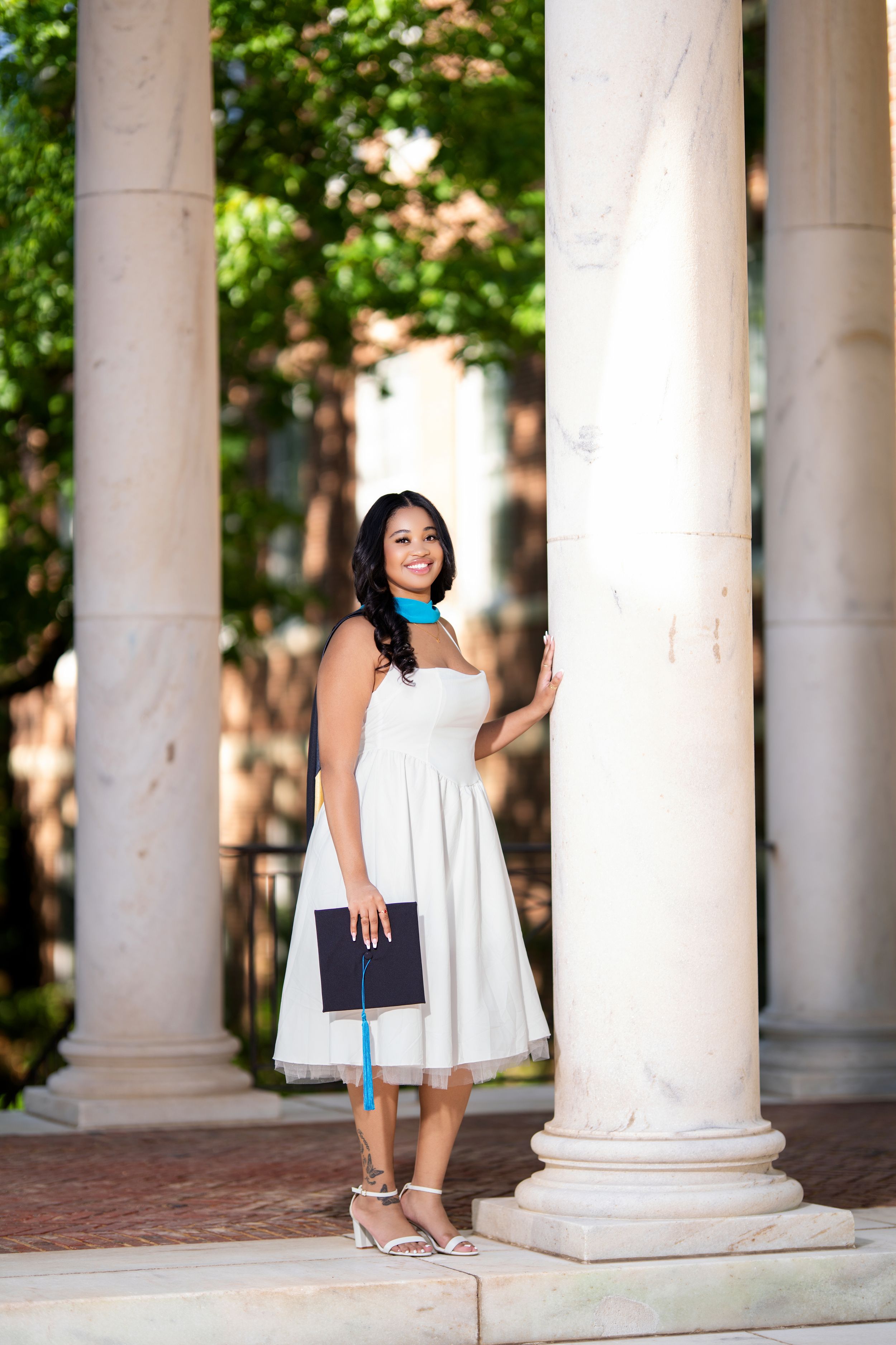 Young woman in white strapless dress holds graduation cap beside white columns at university campus.