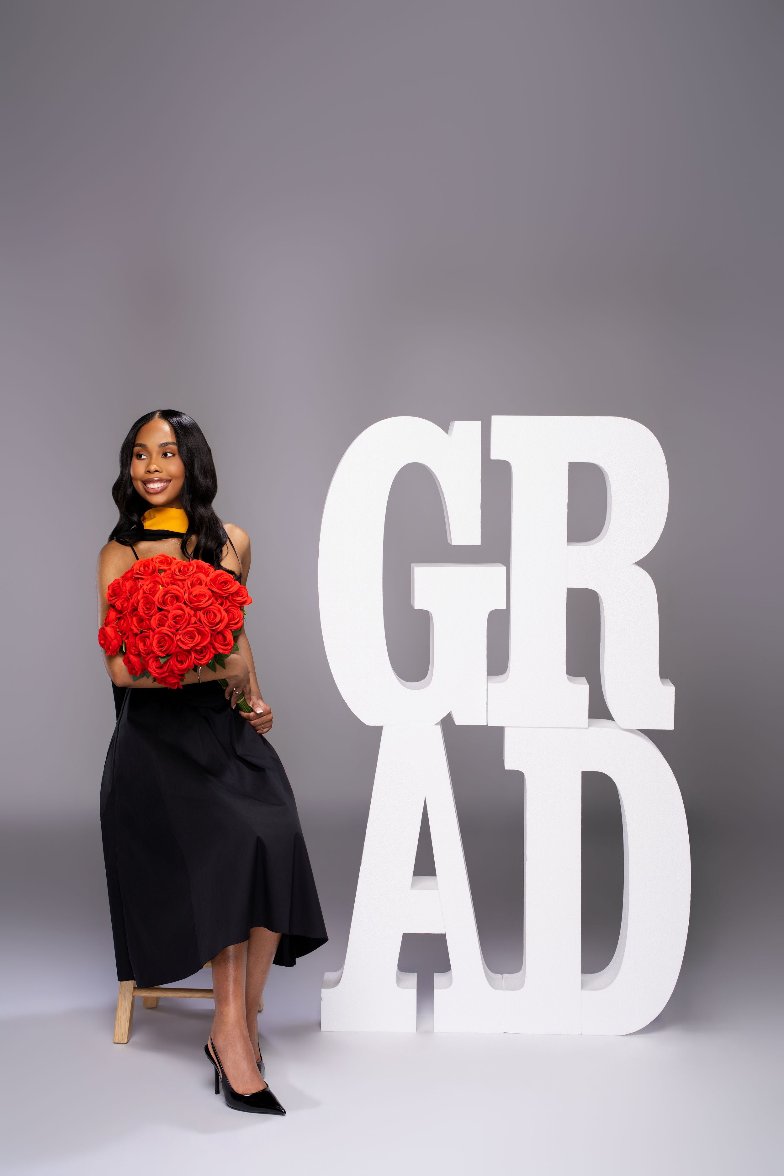 Smiling graduate in black dress holds red roses bouquet beside large white GRAD letters on gray background.