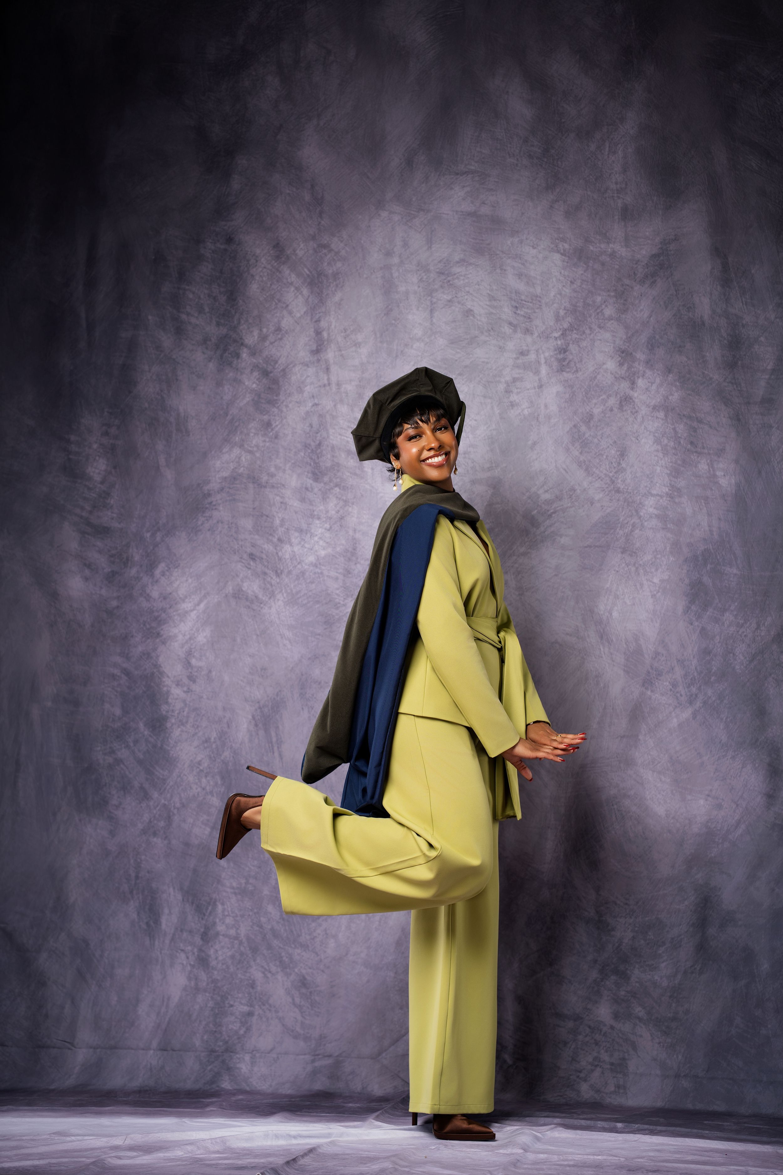 Smiling woman in yellow-green pantsuit and beret poses playfully against textured gray studio background.