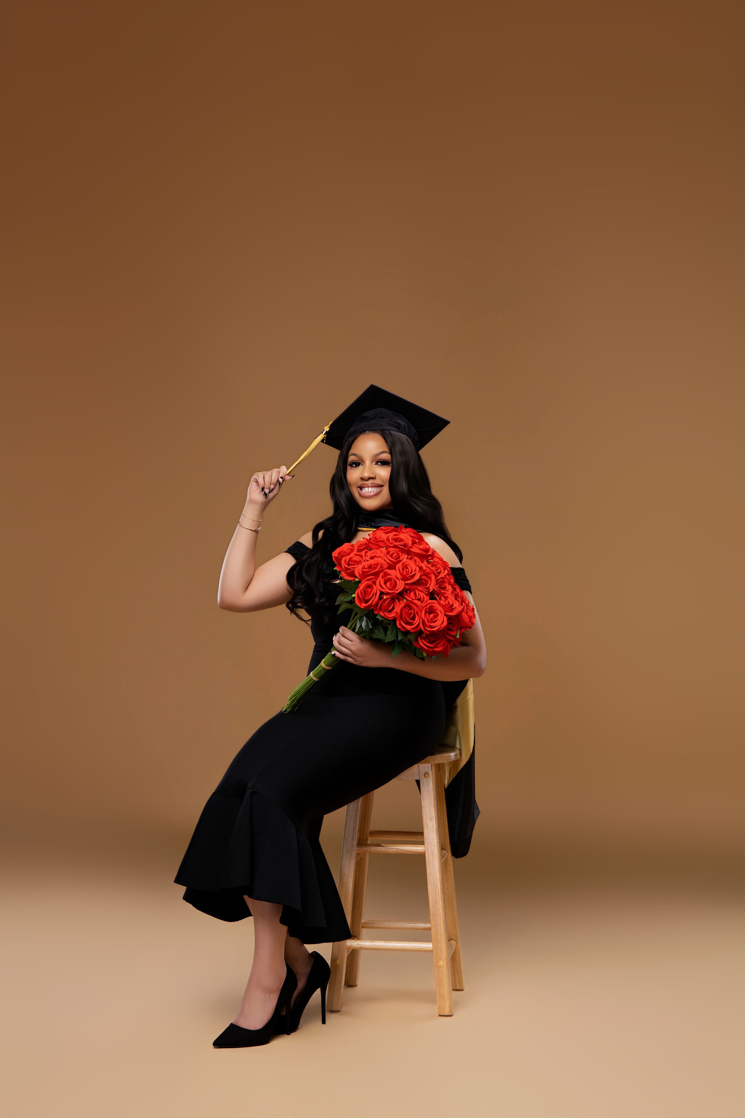 Black woman in black gown and graduation cap holds red roses bouquet while seated on wooden stool.