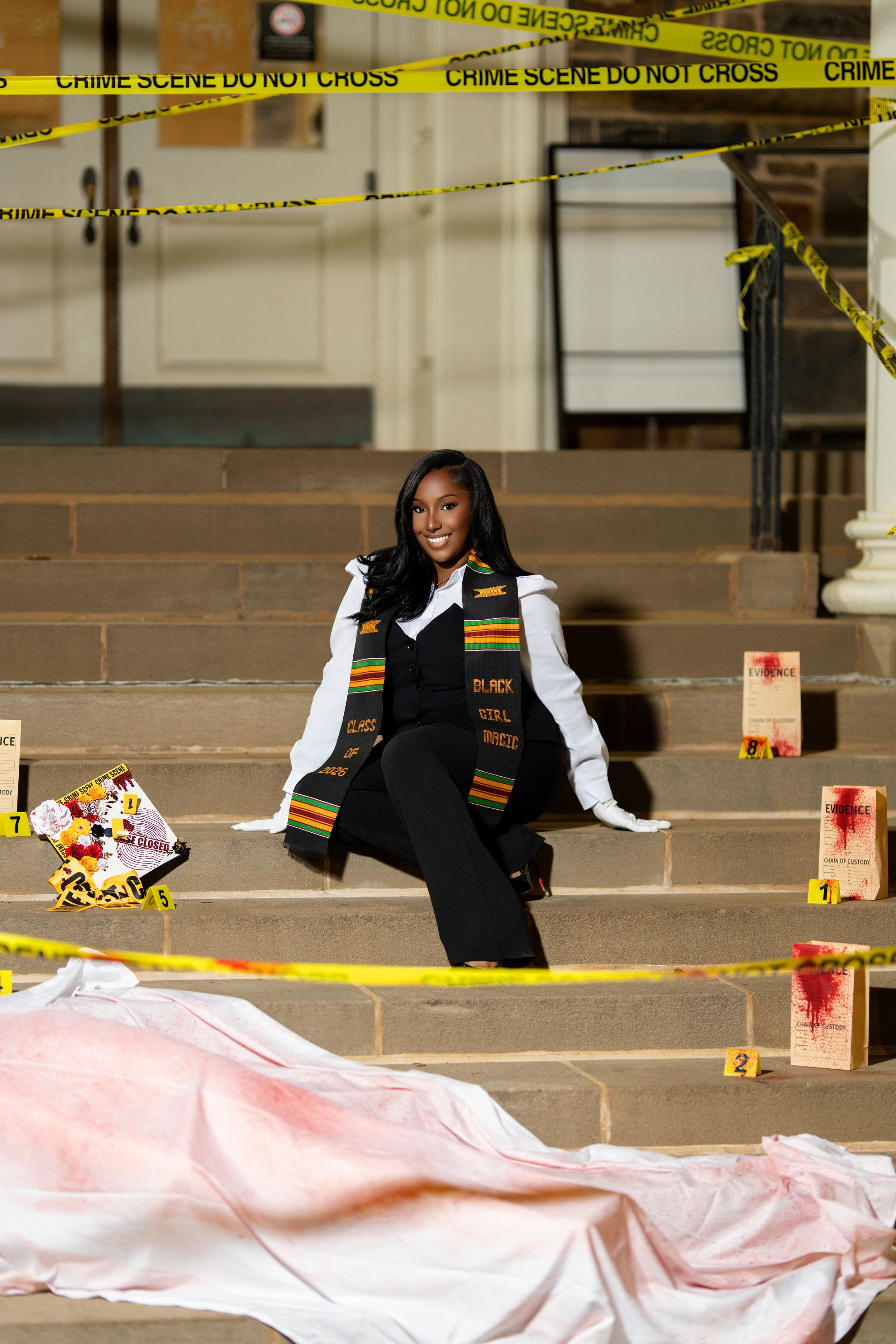 Graduate in kente stole poses on steps near crime scene tape display.