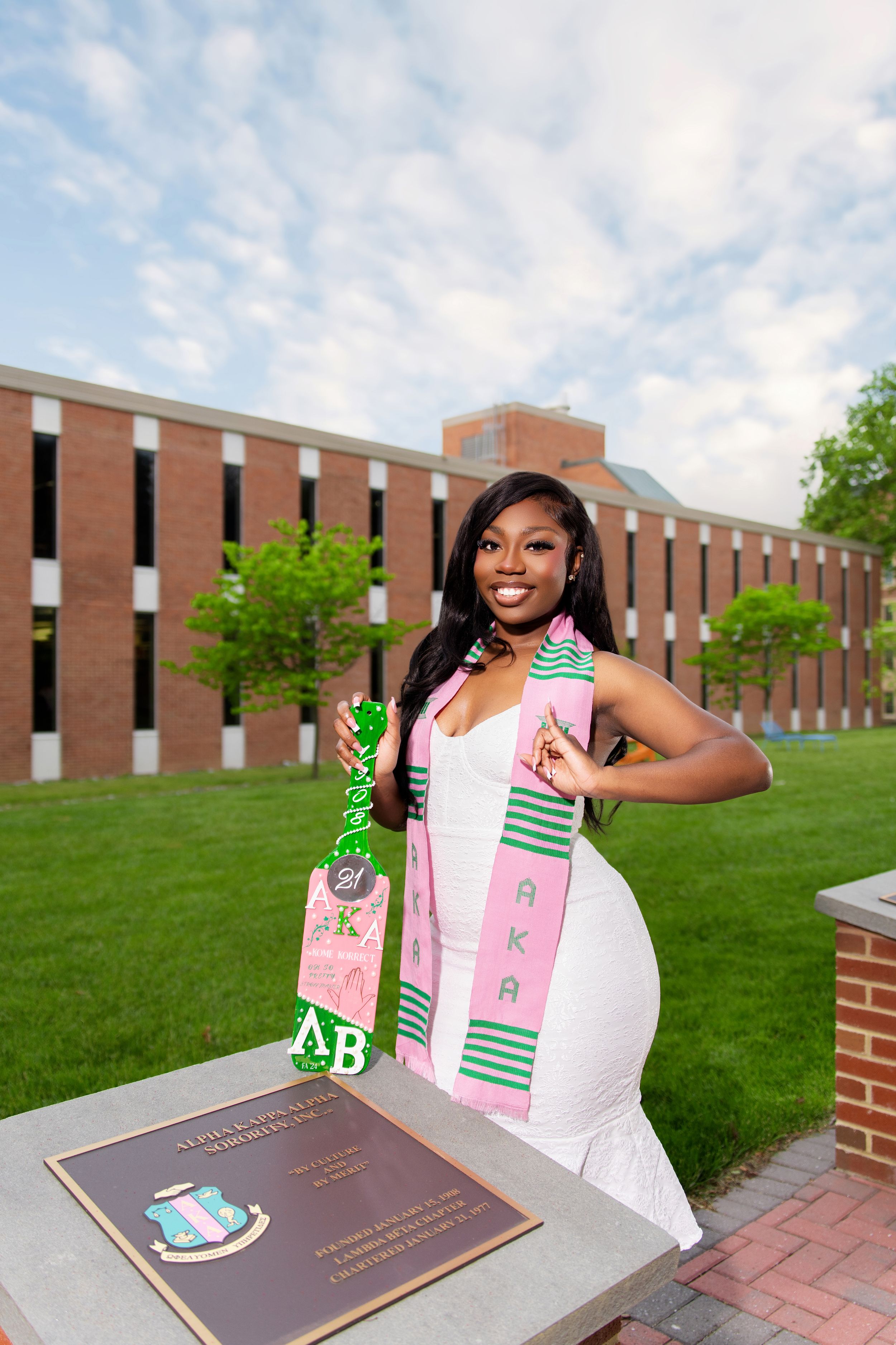 Young woman in white dress poses with AKA sorority paddle and kente sash at college campus graduation photo.