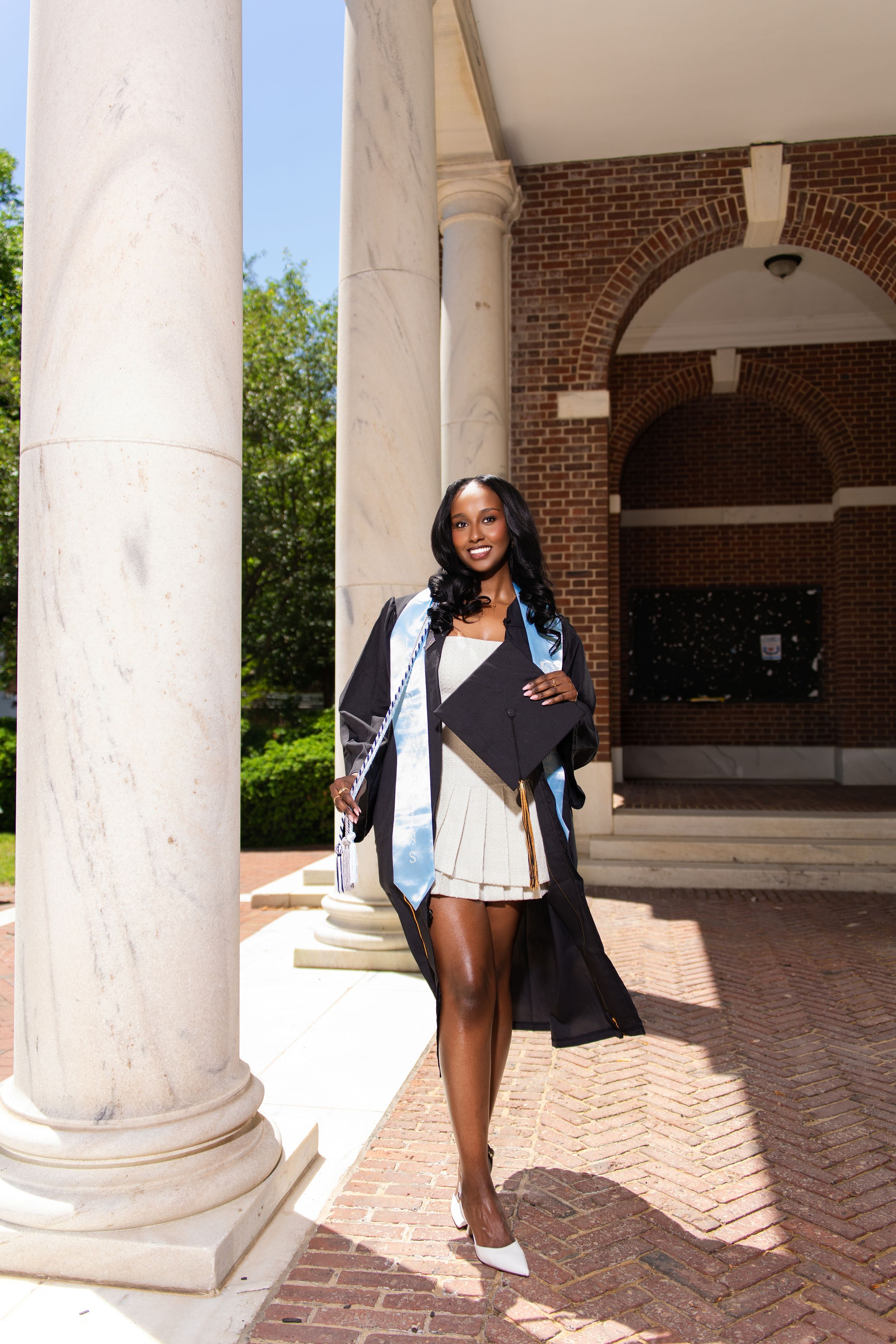 Smiling graduate in cap and gown holds diploma near brick building columns.