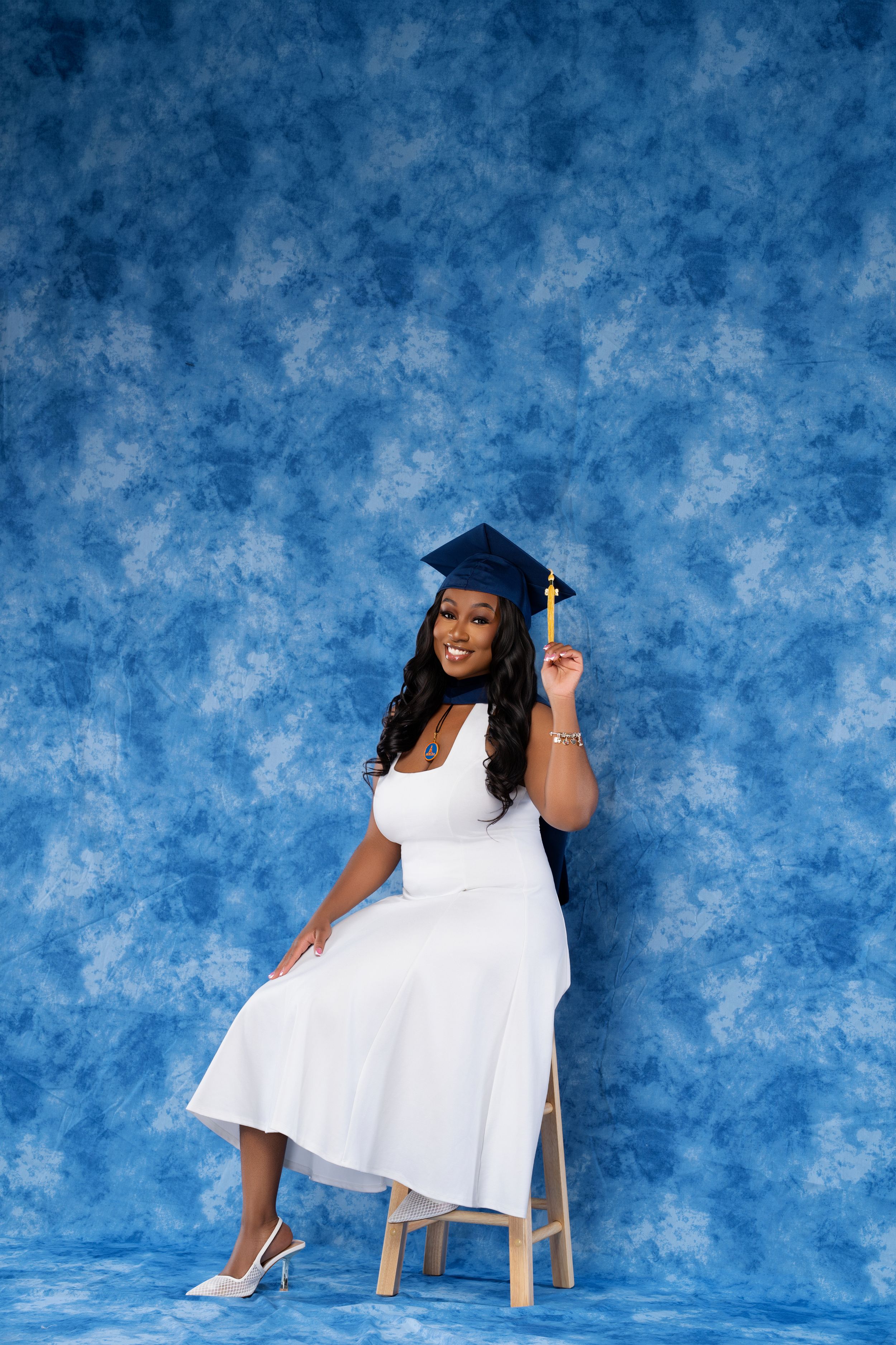 Smiling woman in white dress and blue graduation cap sits on chair against blue mottled backdrop.
