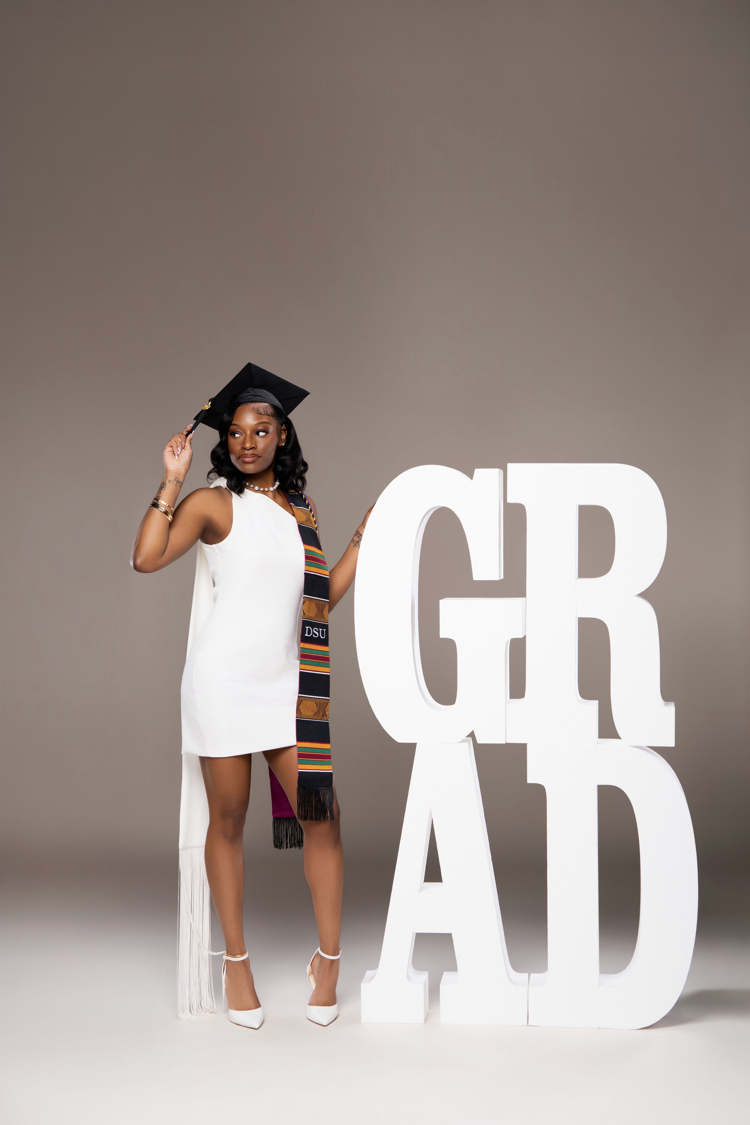 Graduate in white dress and cap poses with kente stole beside large white GRAD letters on gray backdrop.