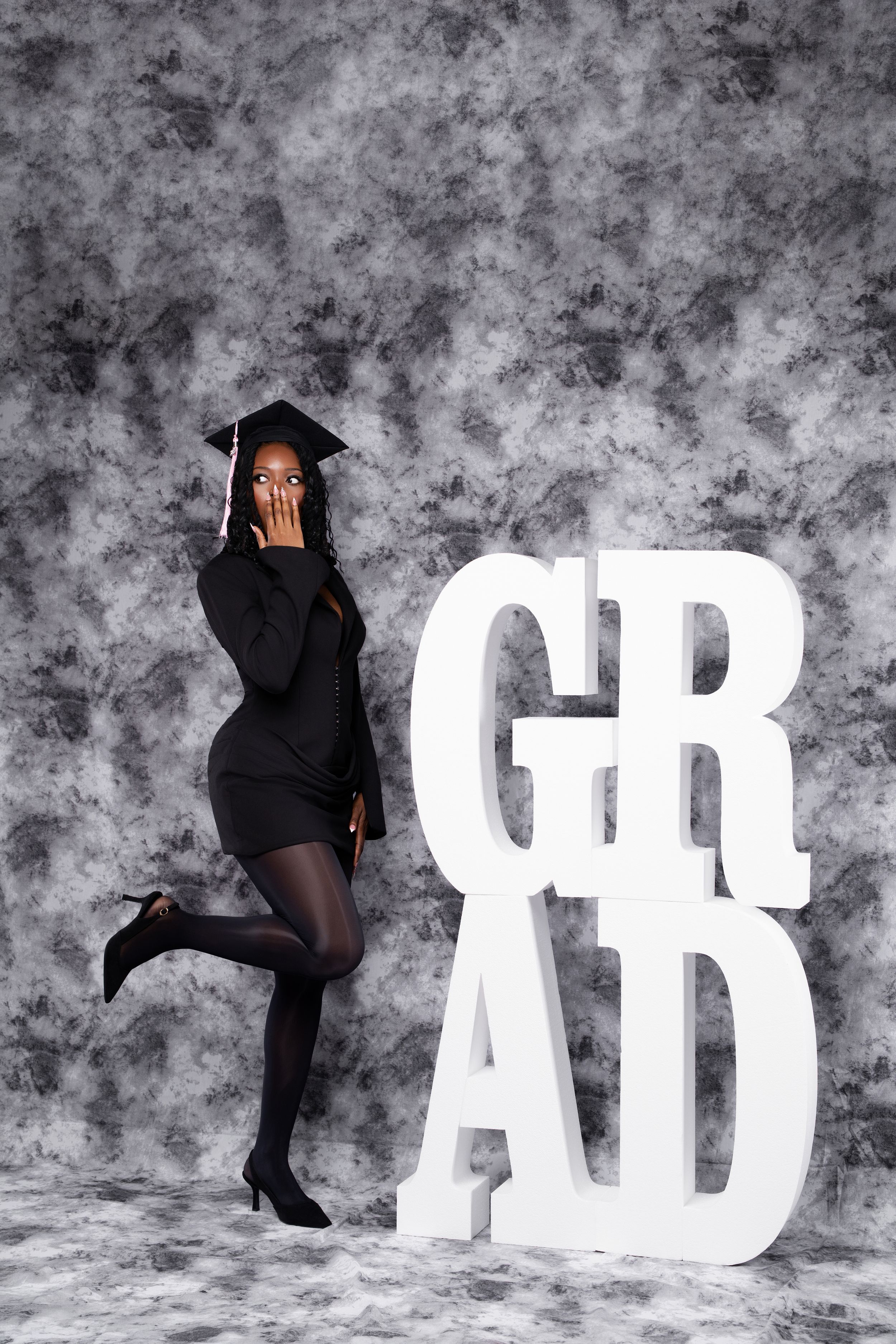 Graduate in black dress and cap poses playfully beside large white GRAD letters on gray backdrop.