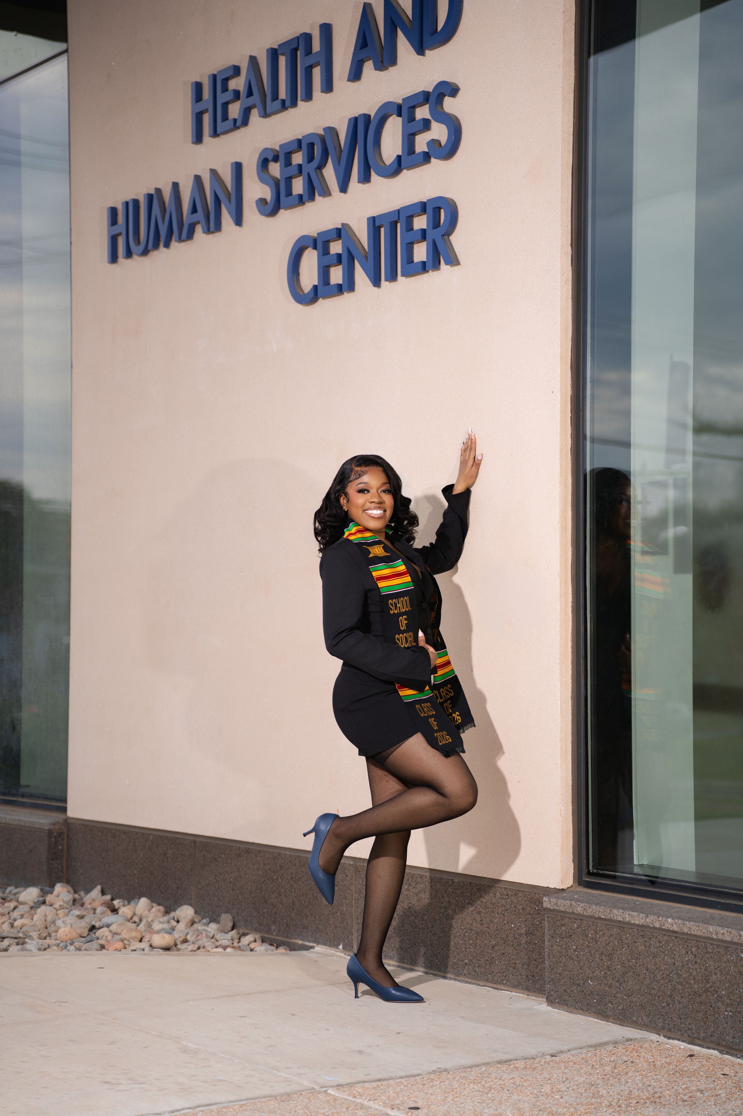 Smiling Black woman in black dress and kente stole poses by Health and Human Services Center sign.