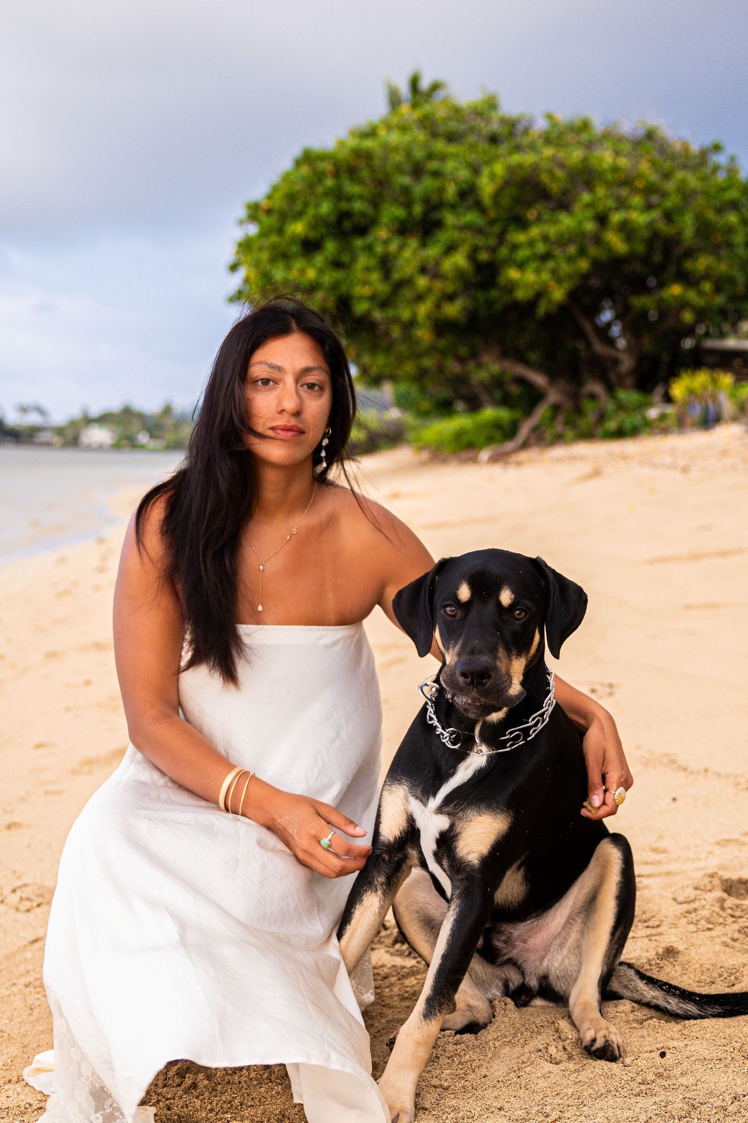 Woman in white dress sitting on beach with black dog, tropical trees and ocean in background.