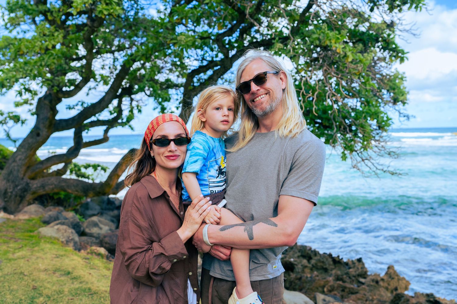 Family of three poses on lush green lawn near rocky Hawaii coastline with ocean views and tropical trees.