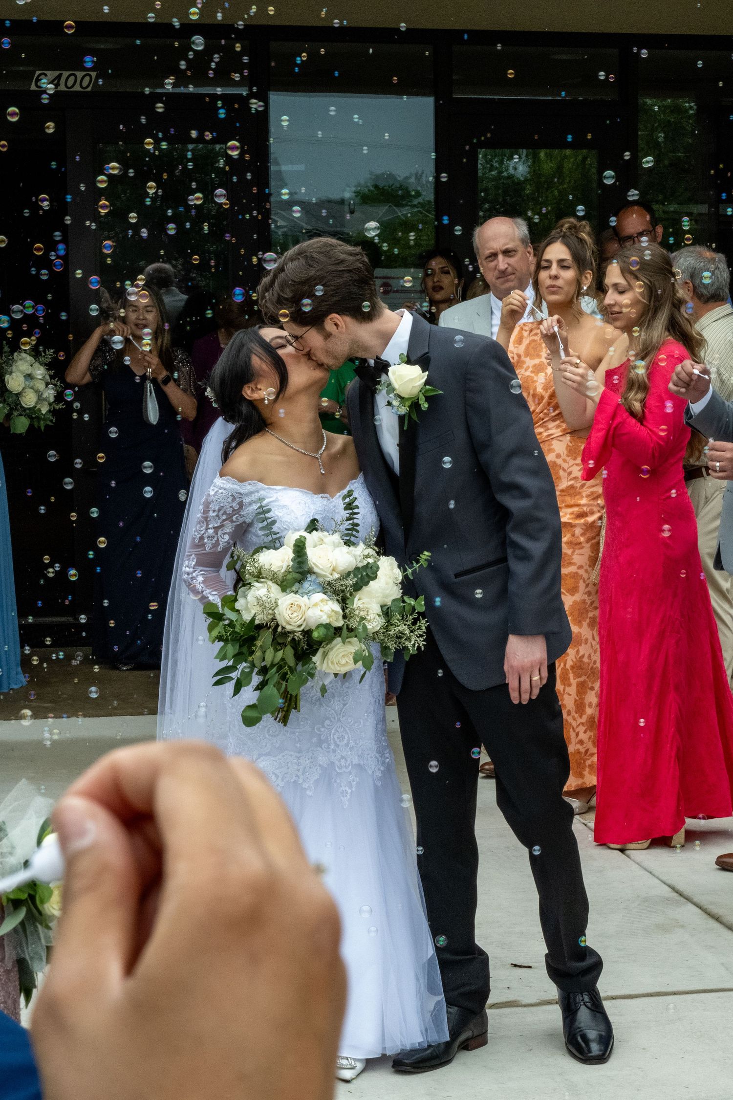 Newlywed couple sharing a kiss surrounded by bubbles and wedding guests outside a venue.