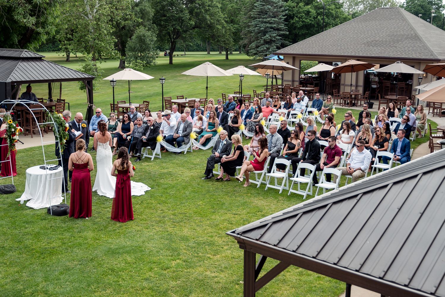 Outdoor wedding ceremony on lush green lawn with guests seated in white chairs near a pavilion on a sunny day.