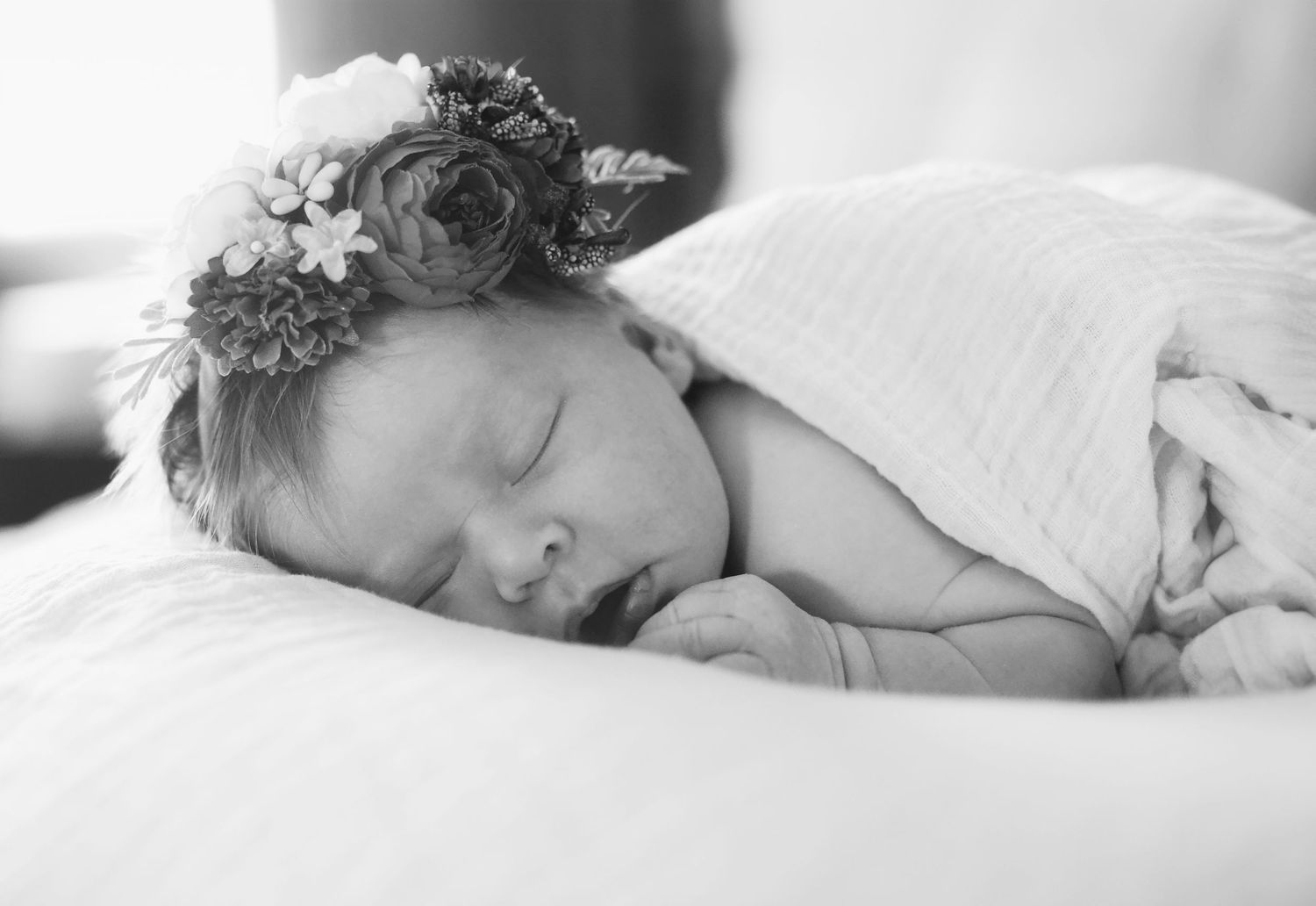 Black and white photo of a sleeping newborn baby girl wearing a delicate floral crown, wrapped in a soft white blanket.