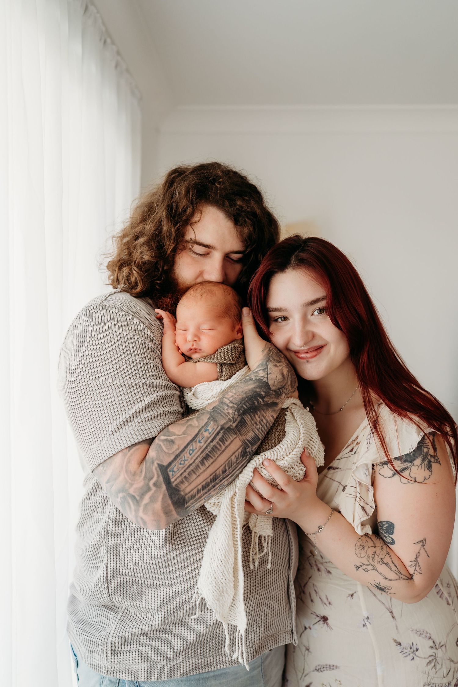 Tattooed couple smiling and holding their sleeping newborn baby wrapped in a white blanket near a bright window.