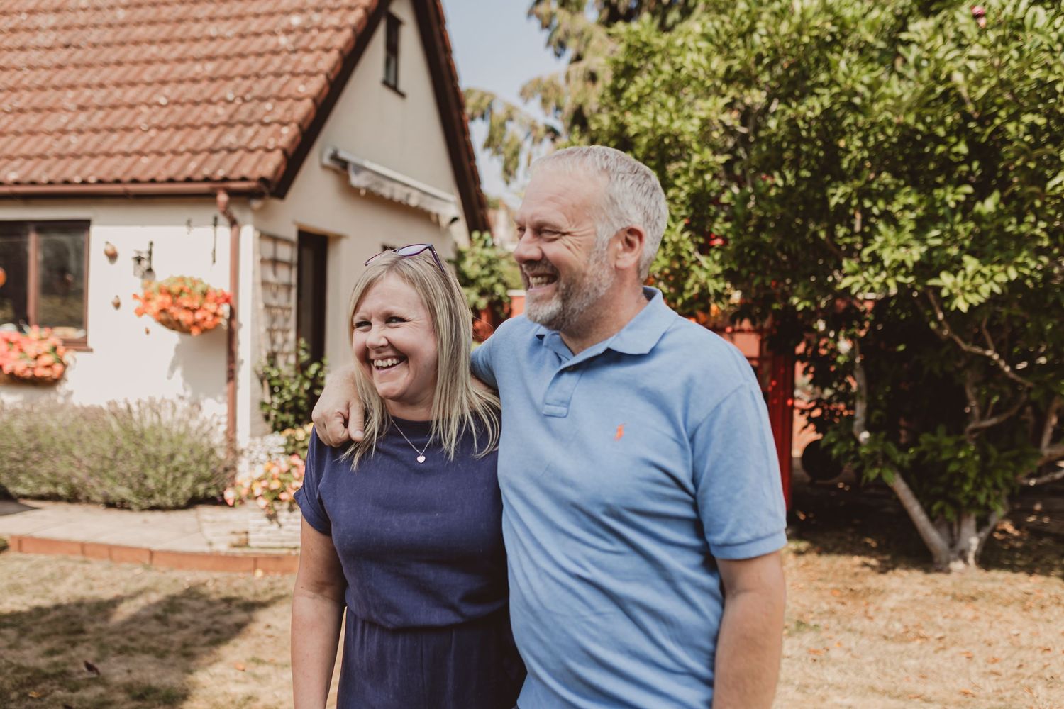 Smiling couple standing together outdoors in front of a cottage garden on a sunny day.