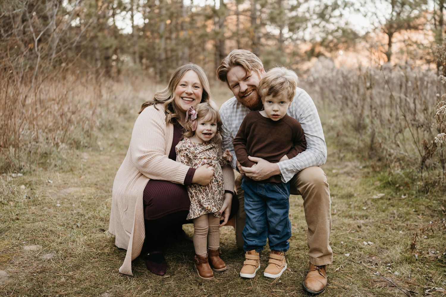 A smiling family of four poses together on a woodland trail at sunset.