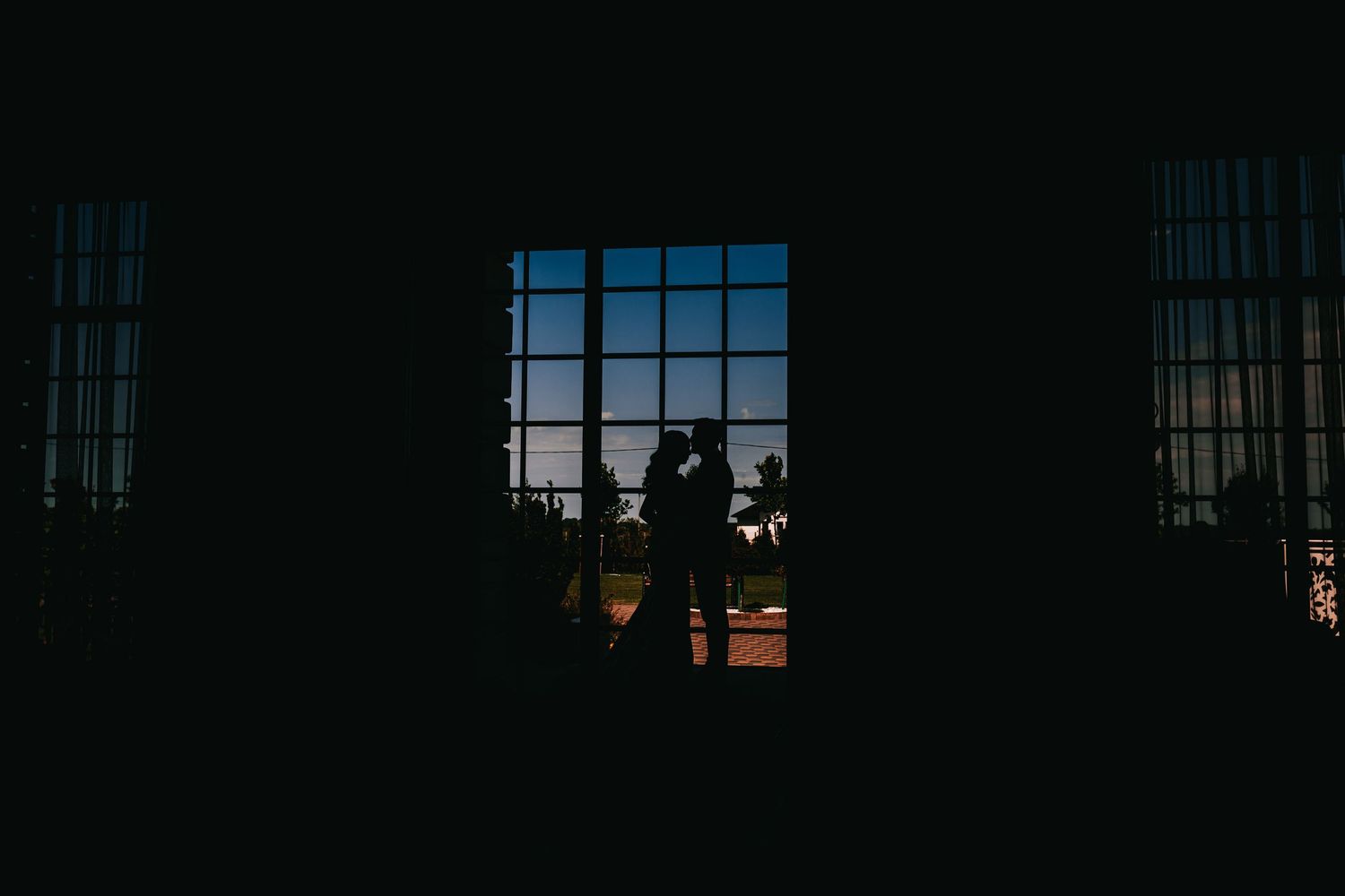 Silhouette of a couple embracing in front of a large window at dusk, creating a romantic and dramatic contrast.