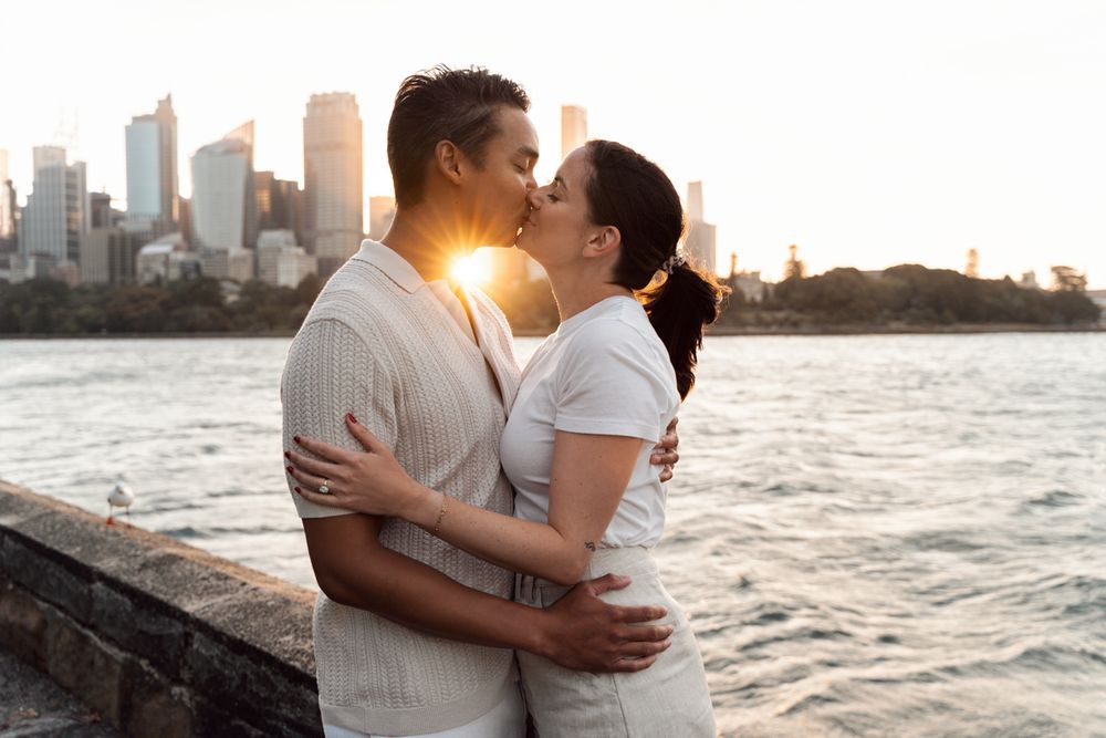 Couple sharing a romantic kiss by the waterfront with a stunning city skyline and golden sunset backdrop. proposal photoshoot sydney