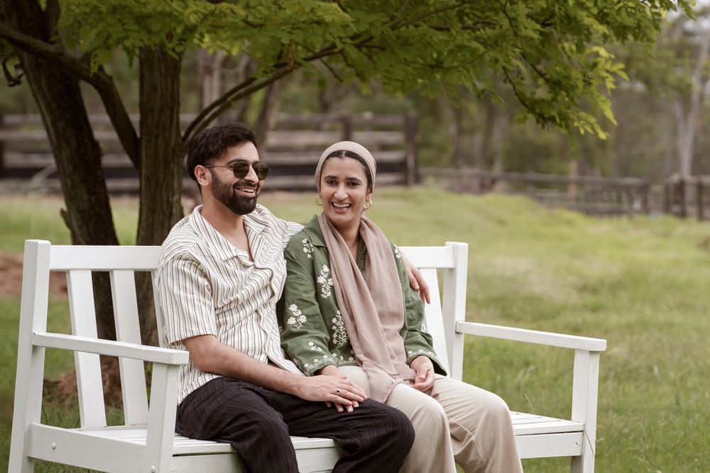 A couple sitting together on a white bench outdoors, smiling, surrounded by green trees and a grassy field cattai couples photoshoot