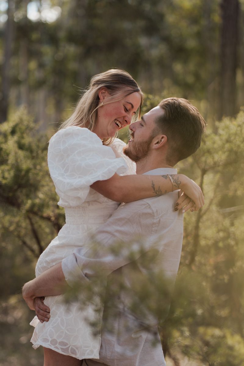 Couple in white outfits embracing and laughing together in a sunlit forest setting proposal photoshoot in rouse hill