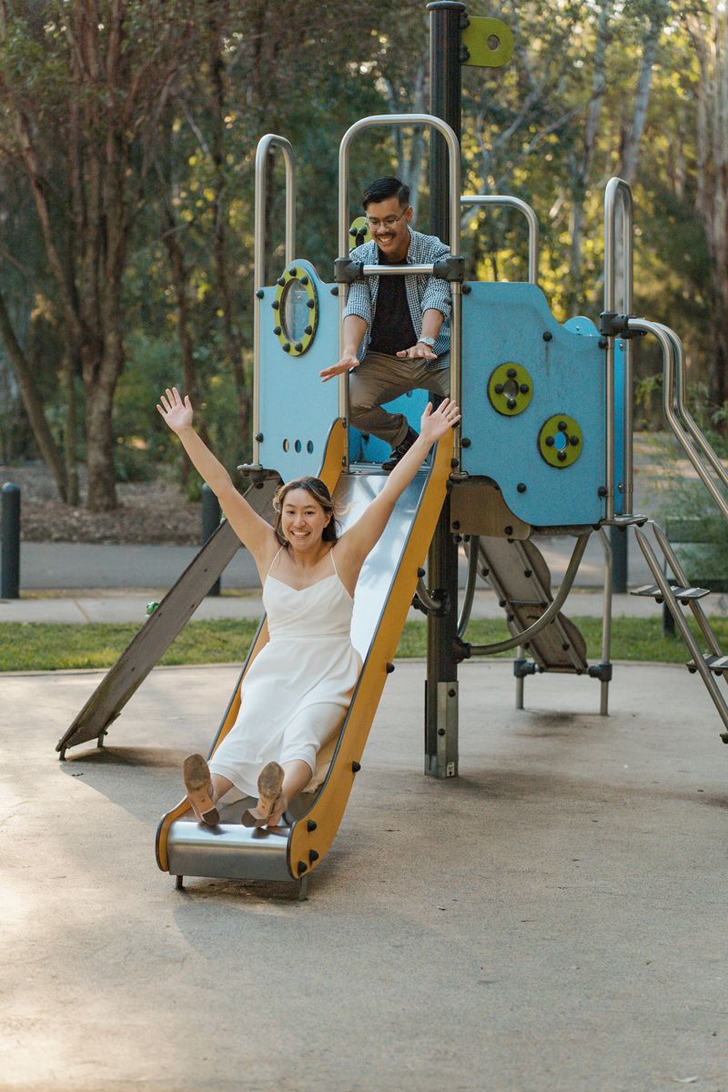 A joyful woman in a white dress slides down a playground slide with arms raised, while a man watches from above fun couples photoshoot