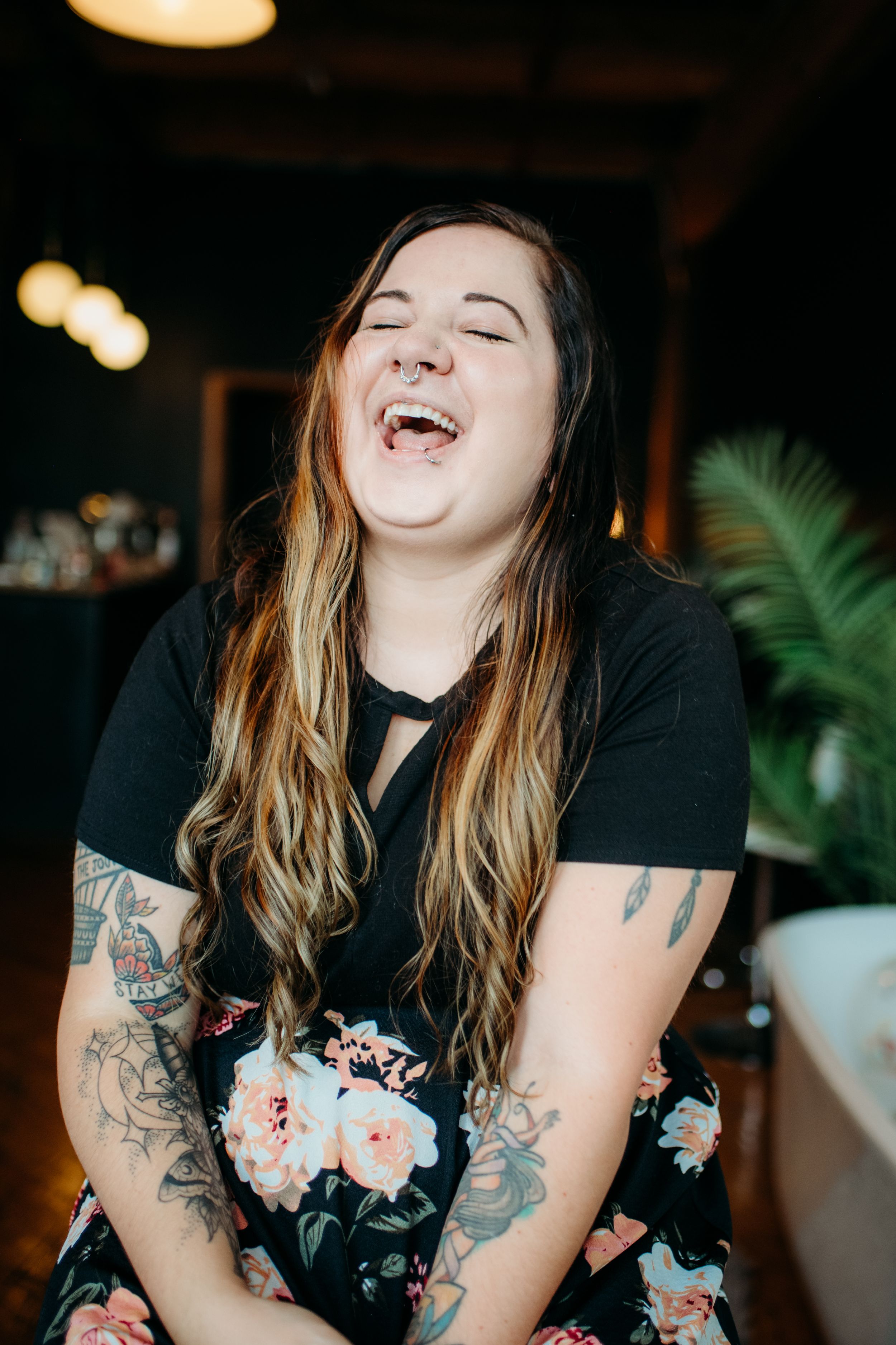 A laughing woman with long hair and tattoos wearing a black floral dress sits in a warmly lit room.