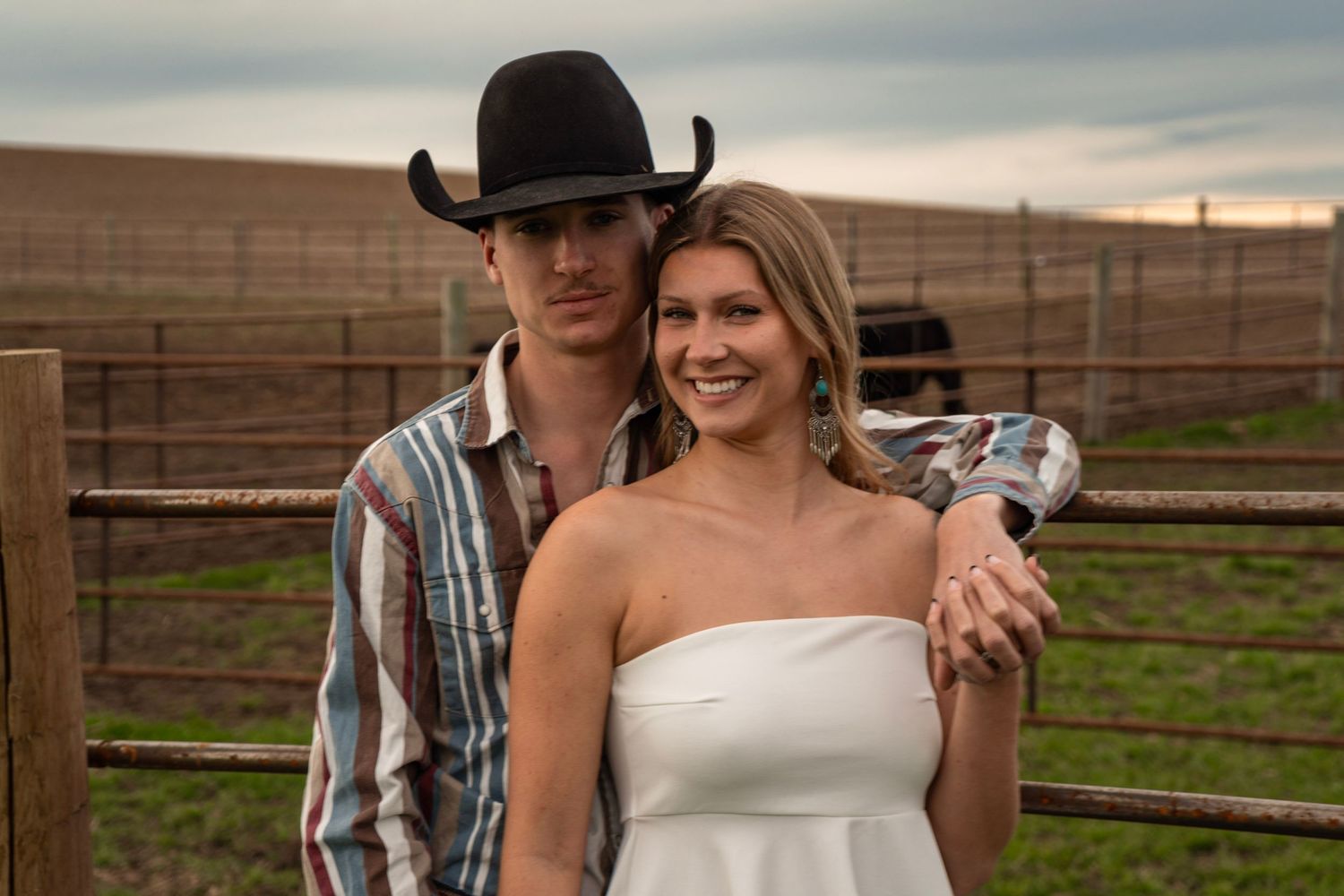 Couple posing at a ranch, man in cowboy hat and plaid shirt with woman in white strapless dress, rustic fence backdrop.