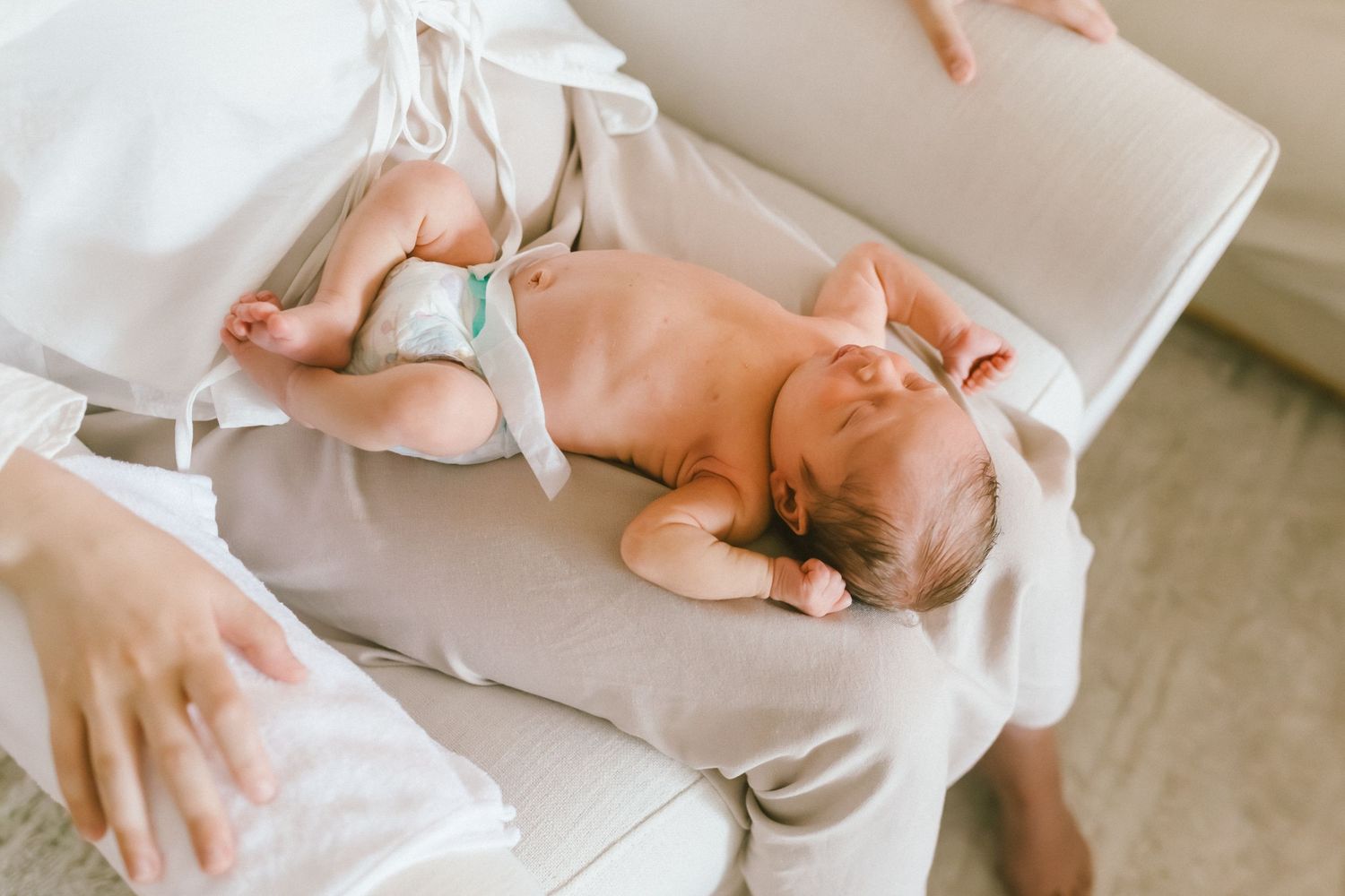 Newborn baby in diaper resting peacefully on mother's lap in soft natural light.
