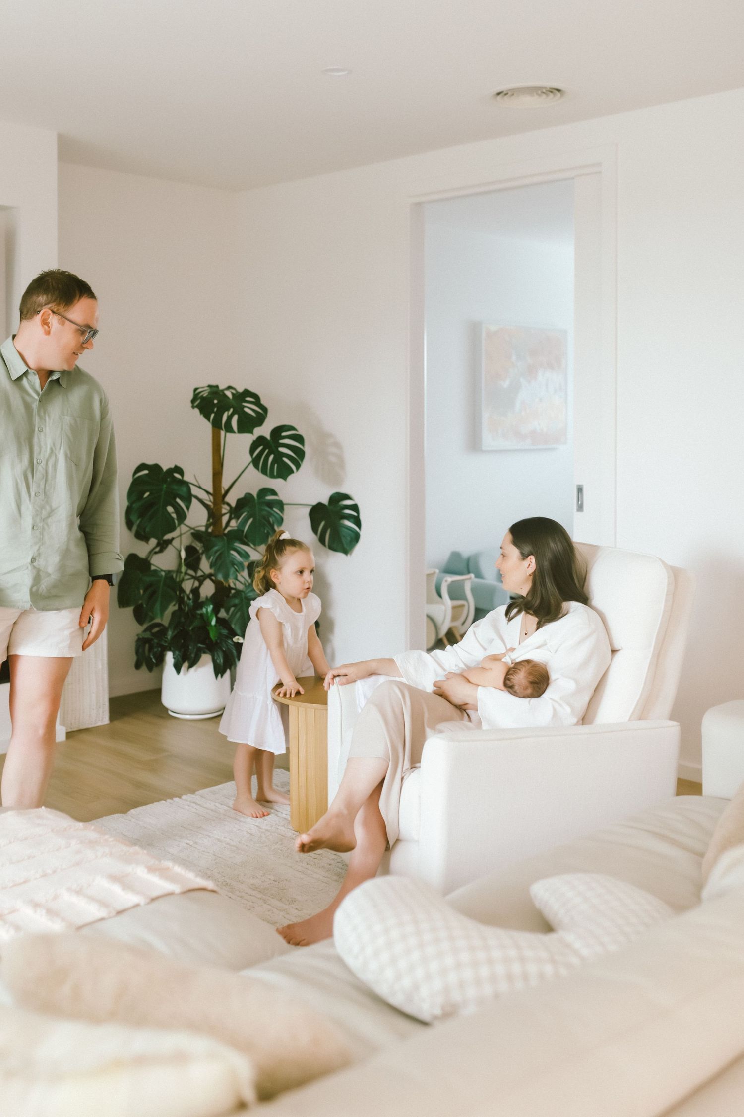 Family at home with mother nursing newborn in chair, toddler playing, and father standing near monstera plant.