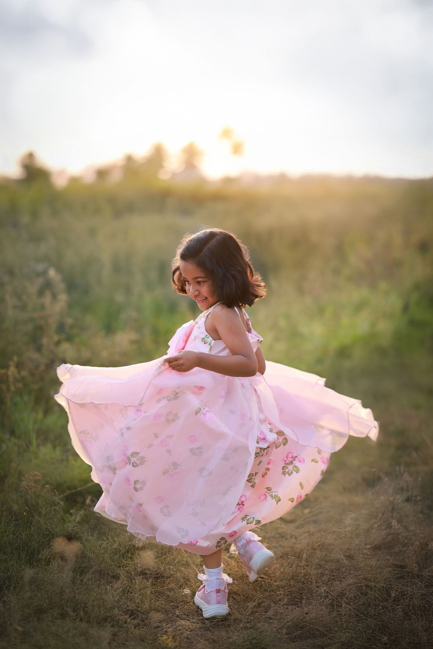A young girl in a pink floral dress twirls joyfully in a sunlit field at golden hour.