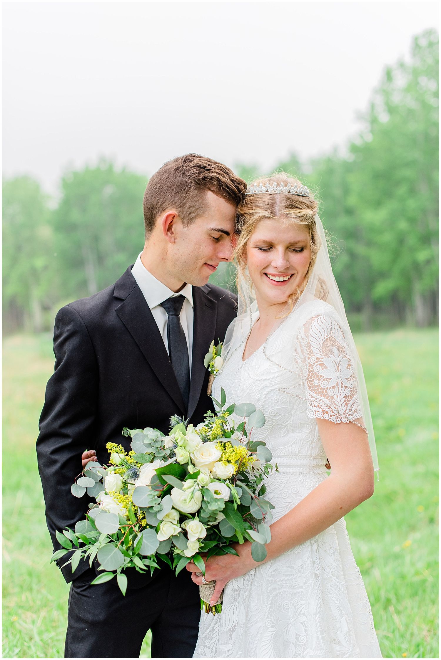 Bride and groom share a tender moment outdoors, bride holding a lush white and green bouquet, wearing lace dress.