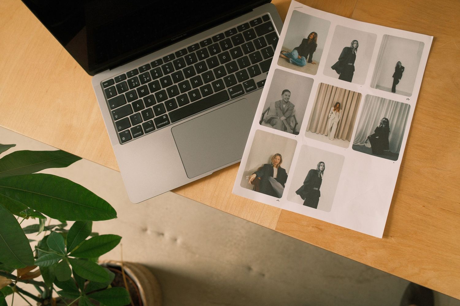 Laptop beside a printed photo contact sheet with fashion portraits on a wooden desk with a green plant.