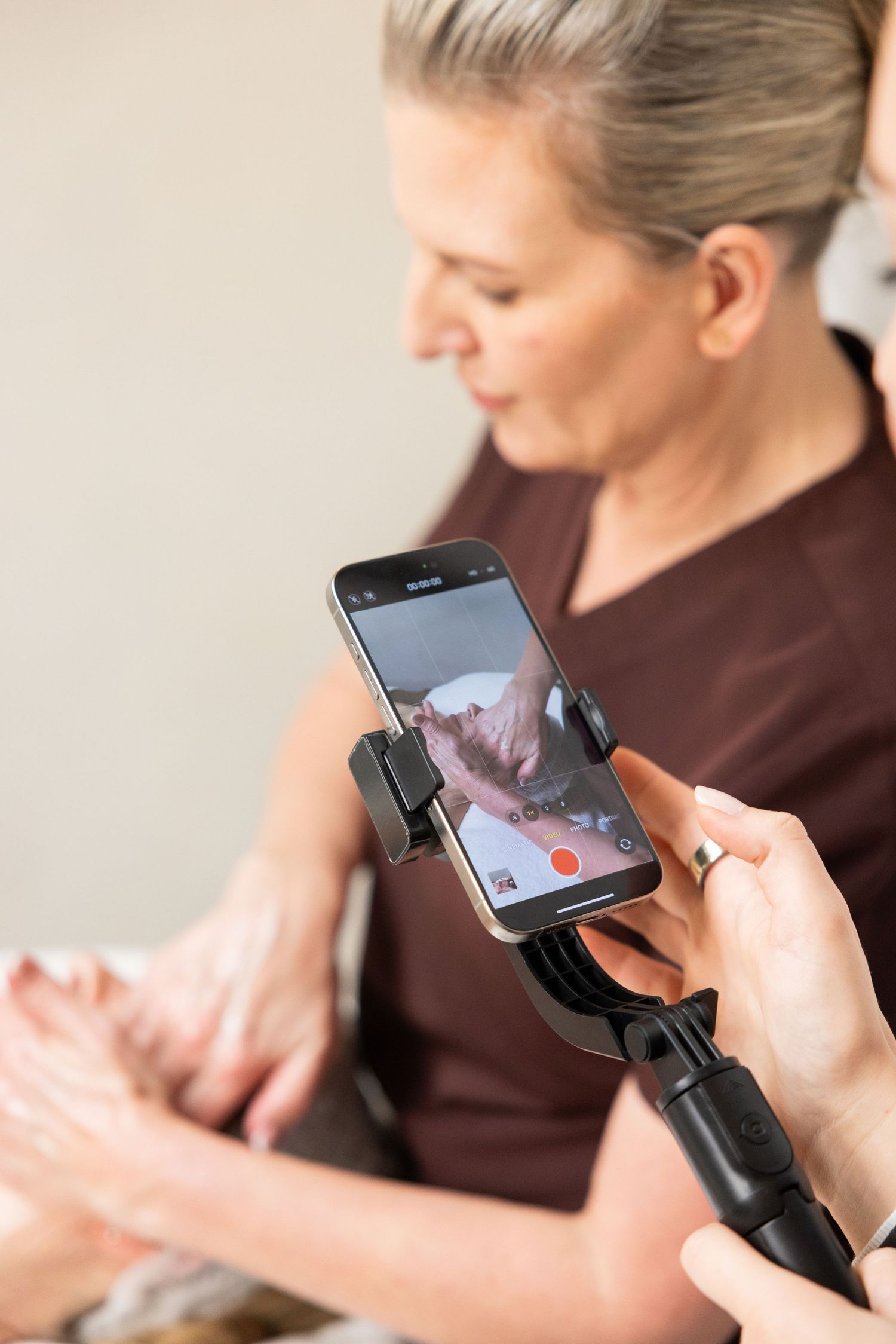 A smartphone on a selfie stick records a massage therapist performing a hand massage on a client in a salon.