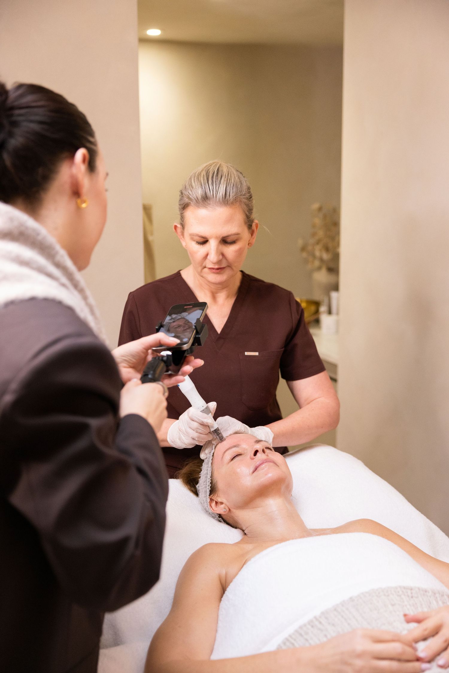 Esthetician performing a facial treatment on a client at a spa while a colleague observes the skincare procedure.