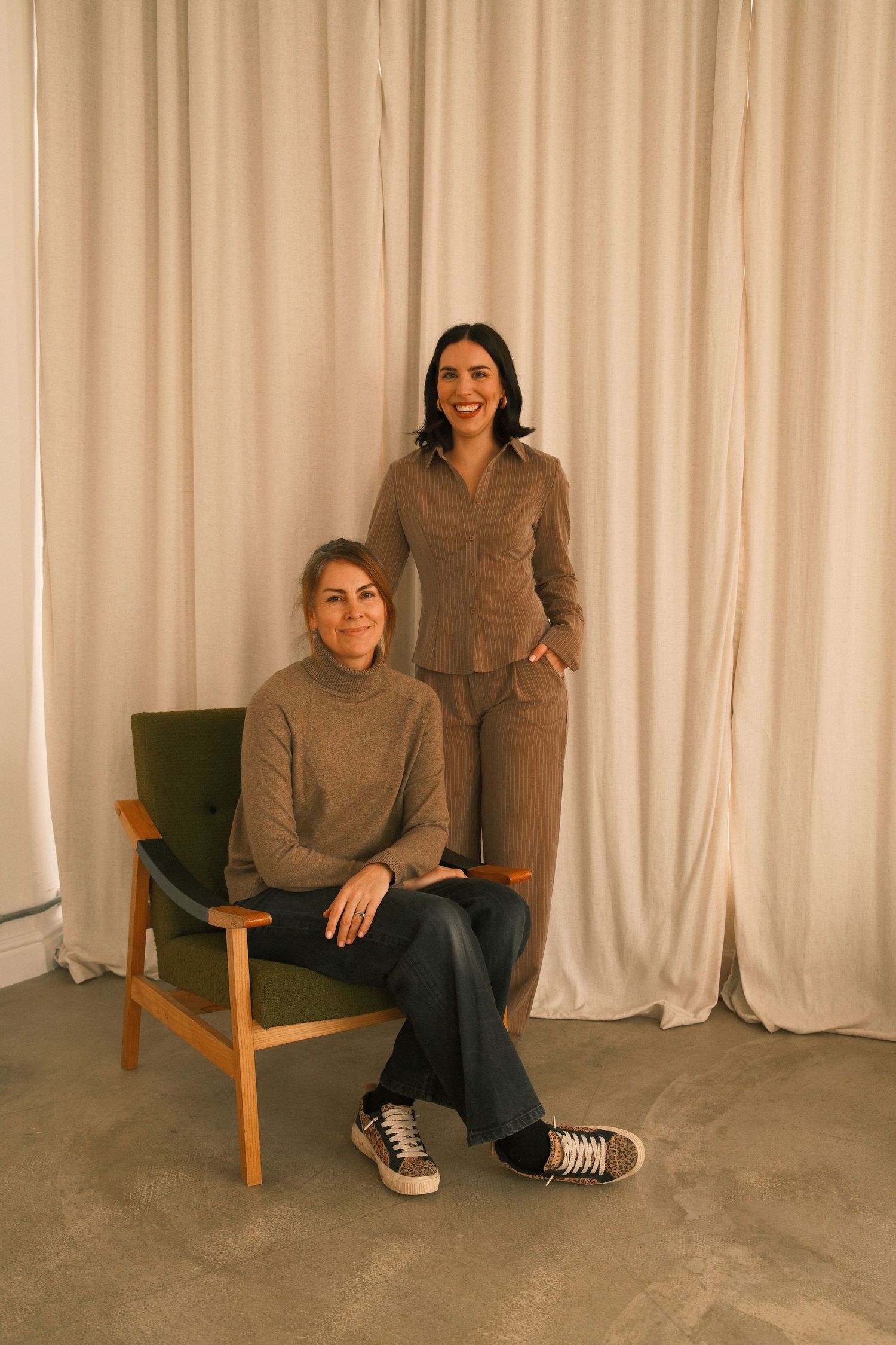 Two women posing in neutral tones, one seated in a green chair, against a white curtain backdrop.