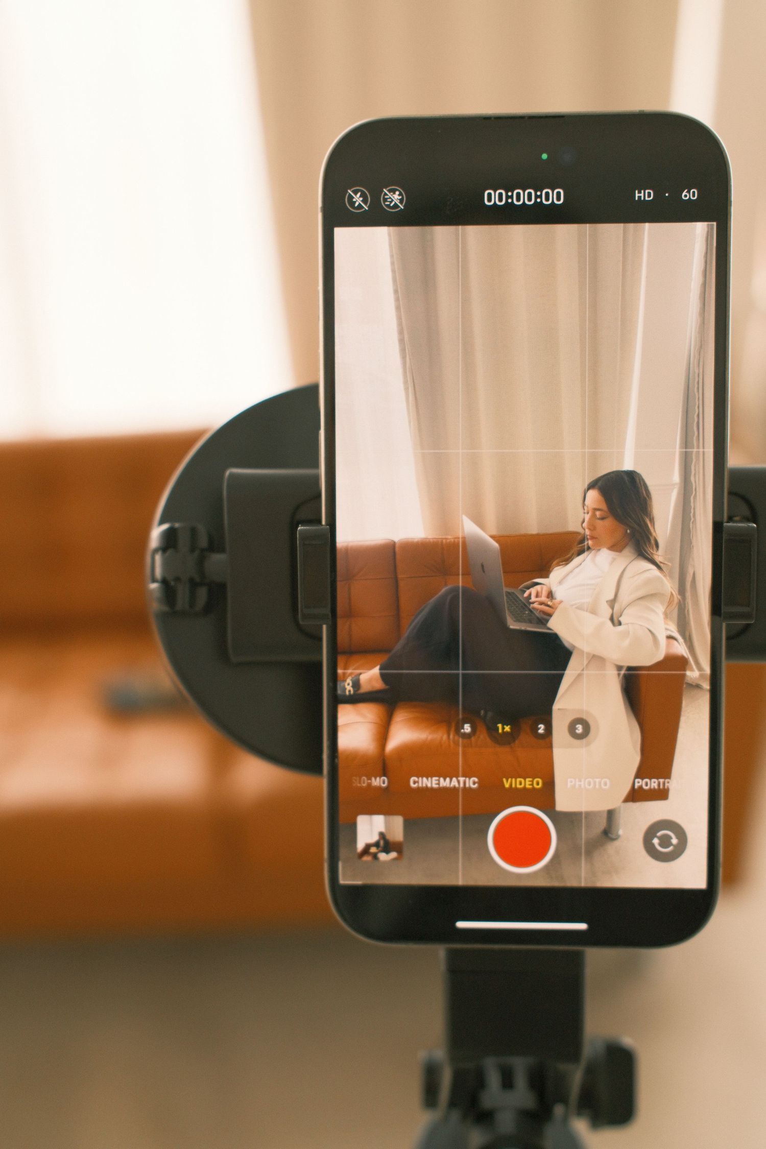 Smartphone on tripod recording a woman sitting on a leather sofa with a laptop in a cozy room.