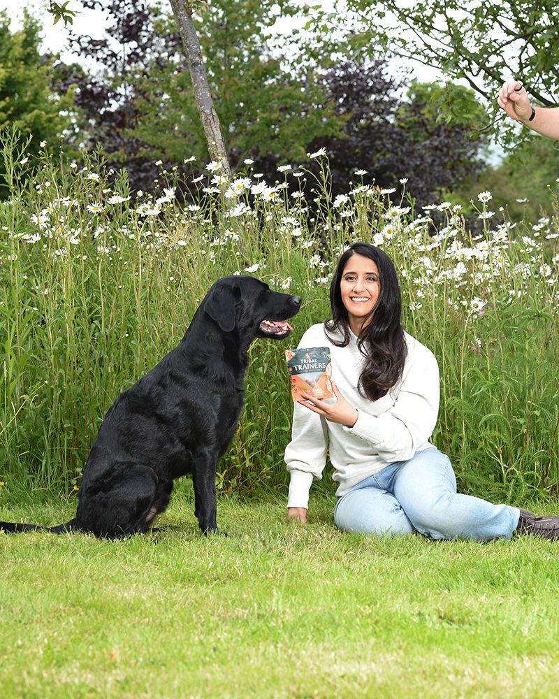 A woman sits on grass holding a pack of dog treats, smiling at a black Labrador. They're in a lush wild meadow garden with blooming daisies.