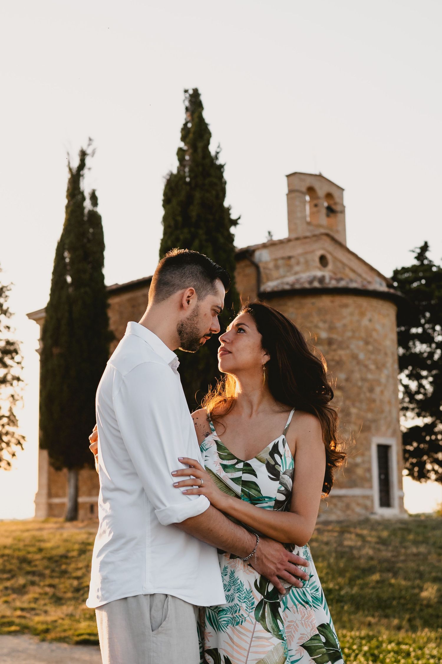 Couple embracing at sunset in front of a rustic stone chapel with cypress trees in Tuscany, Italy.