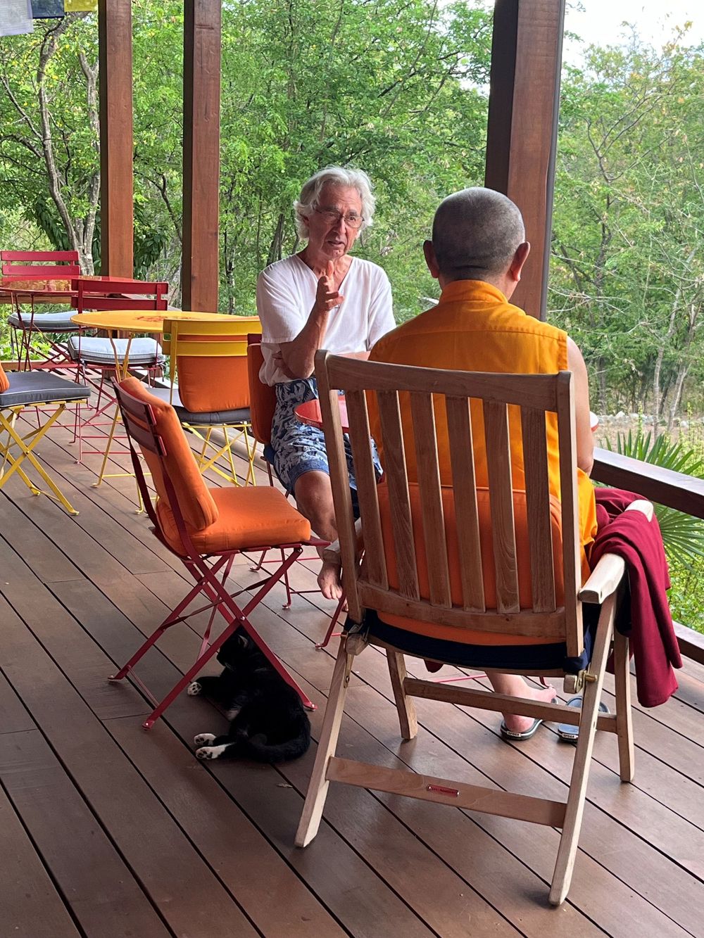 Two people in conversation on a wooden deck, one in white and one in orange robes, surrounded by lush green trees.