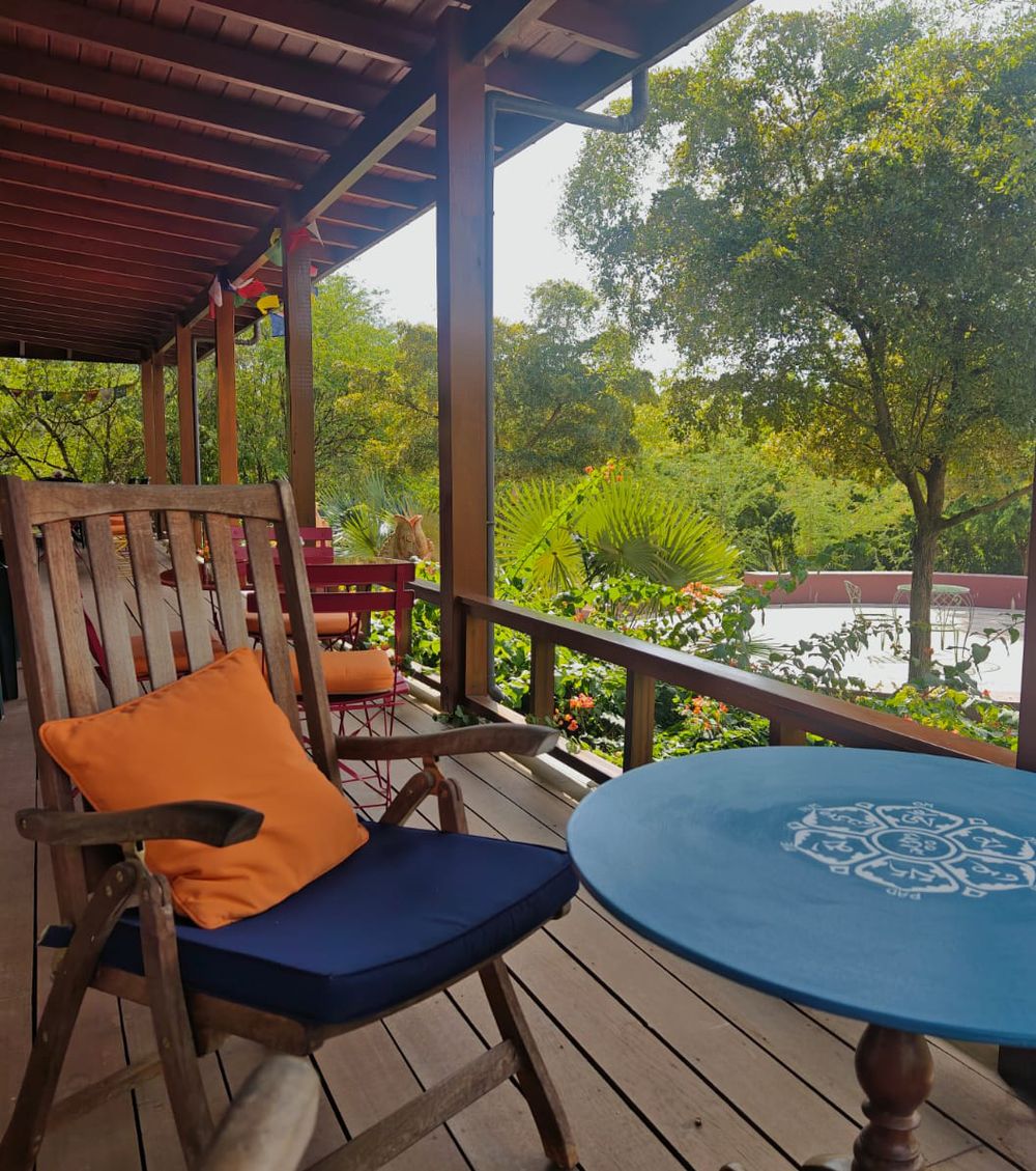 Wooden deck with rocking chair, orange and blue cushions, teal table, overlooking lush tropical garden and pond.