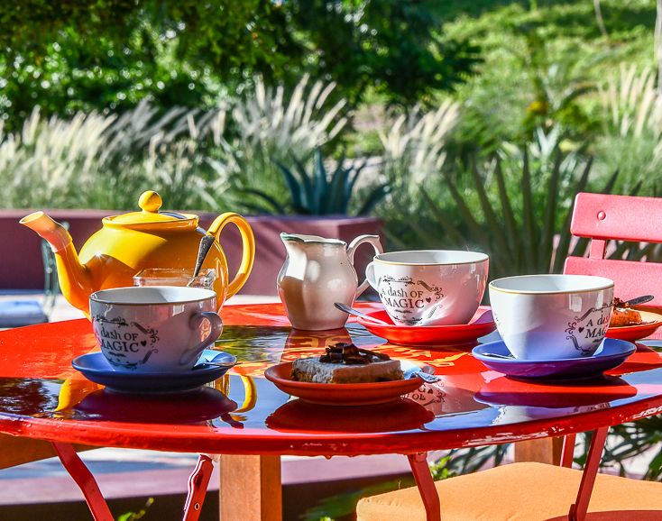 Colorful outdoor tea setting with yellow teapot, cups, and saucers on a red table surrounded by lush greenery.