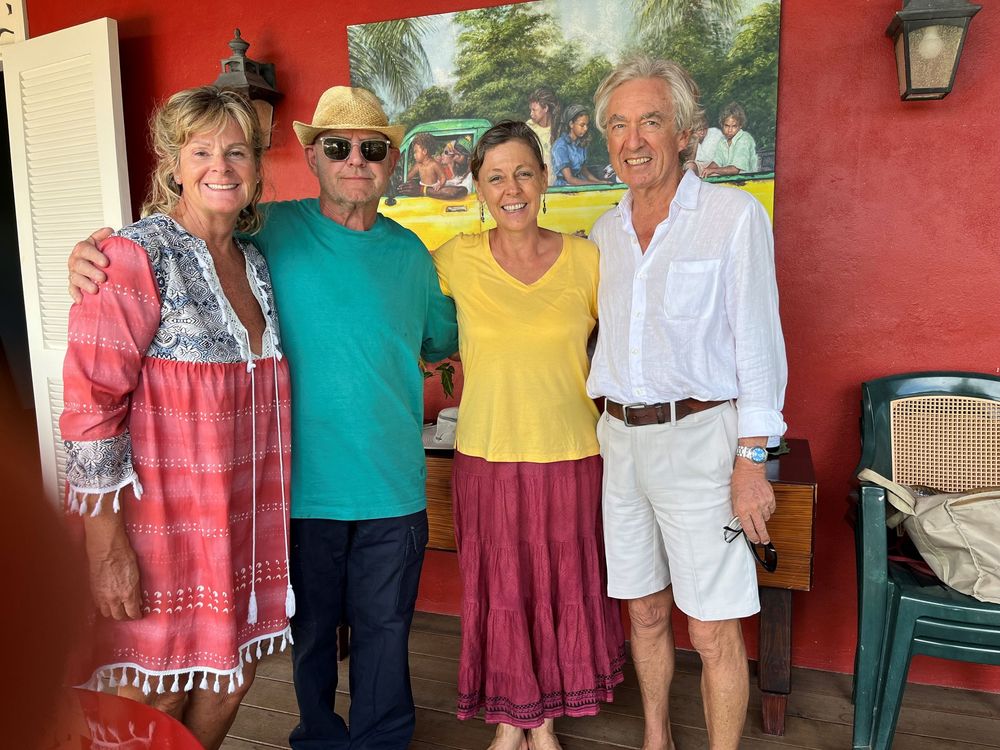 Four smiling friends pose together indoors near a colorful mural, dressed in casual vacation attire.