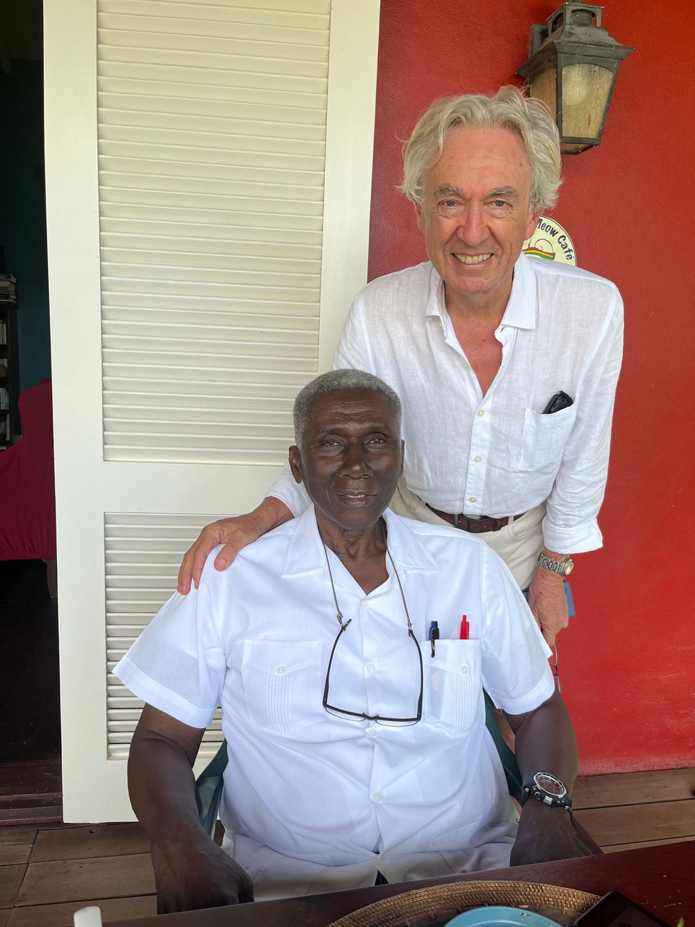 Two men posing together on a colorful porch, one seated wearing white shirt with pens, other standing in linen shirt smiling.