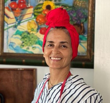Smiling woman wearing a red turban and striped shirt stands in front of a colorful fruit painting.