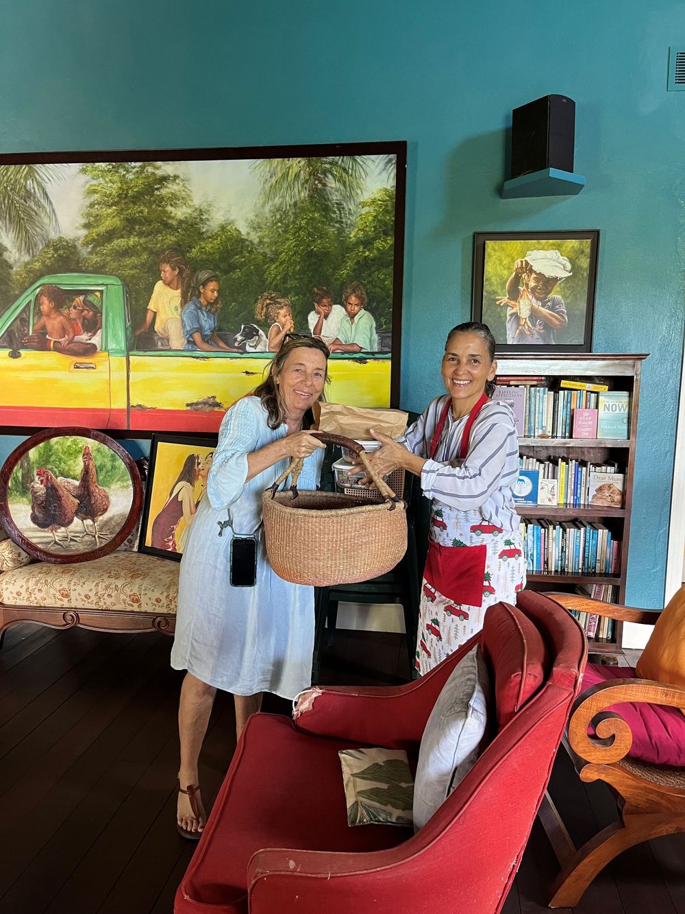 Two women smiling and holding a woven basket together in a colorful room with artwork and bookshelves.