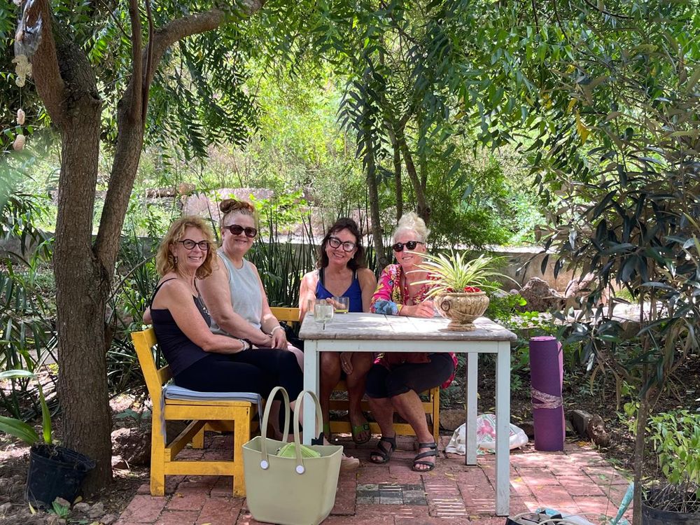 Four women sitting together at a white outdoor table surrounded by lush green trees in a sunny garden setting.
