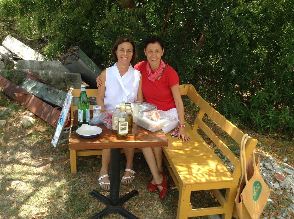 Two women sitting at a yellow wooden table outdoors, with drinks and food, surrounded by lush green trees.